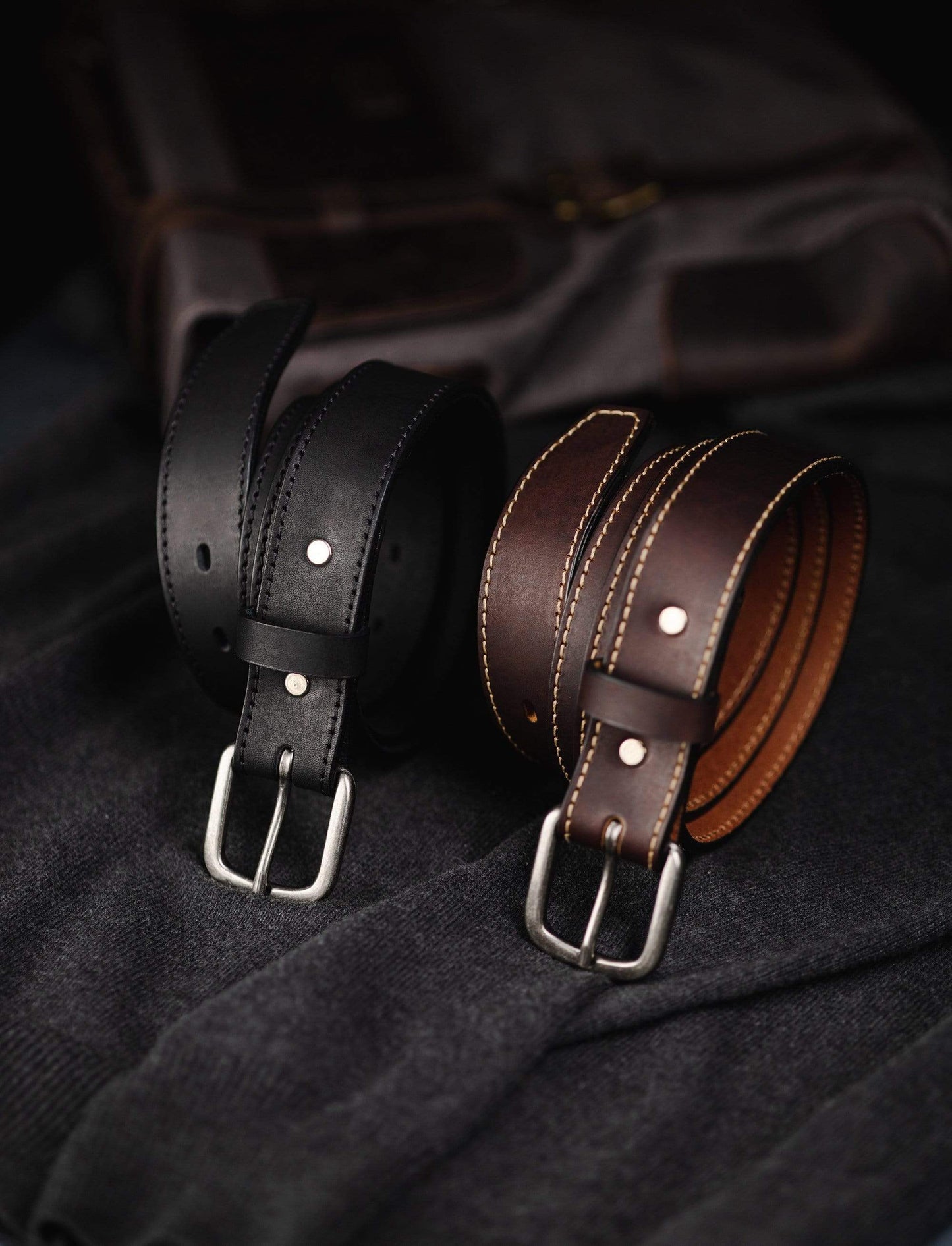 Two leather belts, one black and one brown, on a dark fabric background.