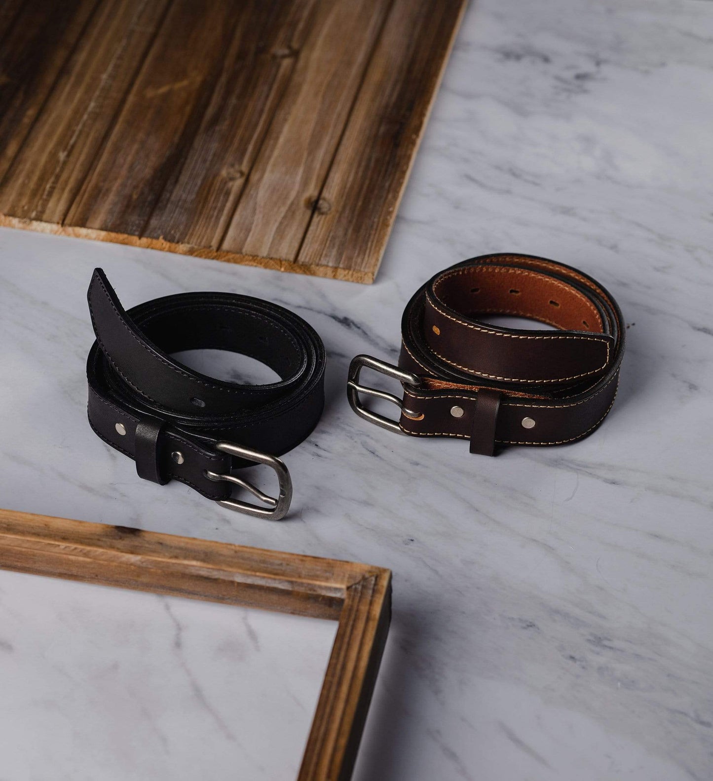 Two leather belts, one black and one brown, on a marble surface with a wooden frame.