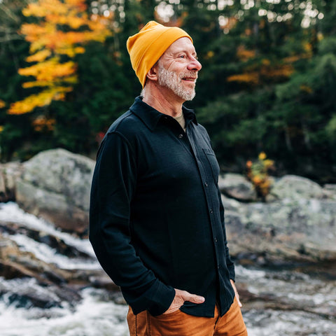 Man wearing a black Merino wool shirt and yellow beanie by a river, surrounded by autumn foliage.