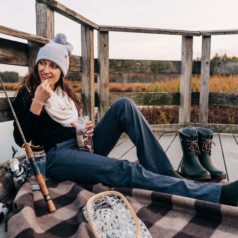 Woman enjoying snacks by a lake in warm clothing, surrounded by fishing gear and cozy blankets.