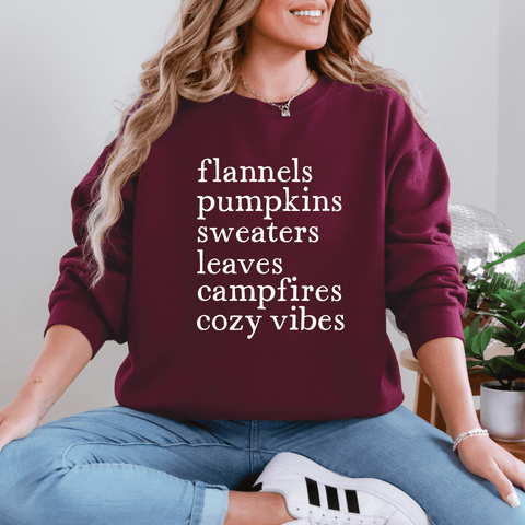 Woman wearing a burgundy sweatshirt with fall-themed words: flannels, pumpkins, sweaters, leaves, campfires, cozy vibes.