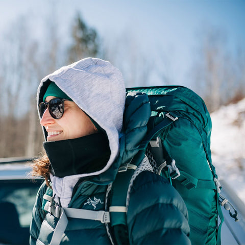 Smiling woman in sunglasses wearing a hooded jacket and carrying a backpack in a snowy landscape.