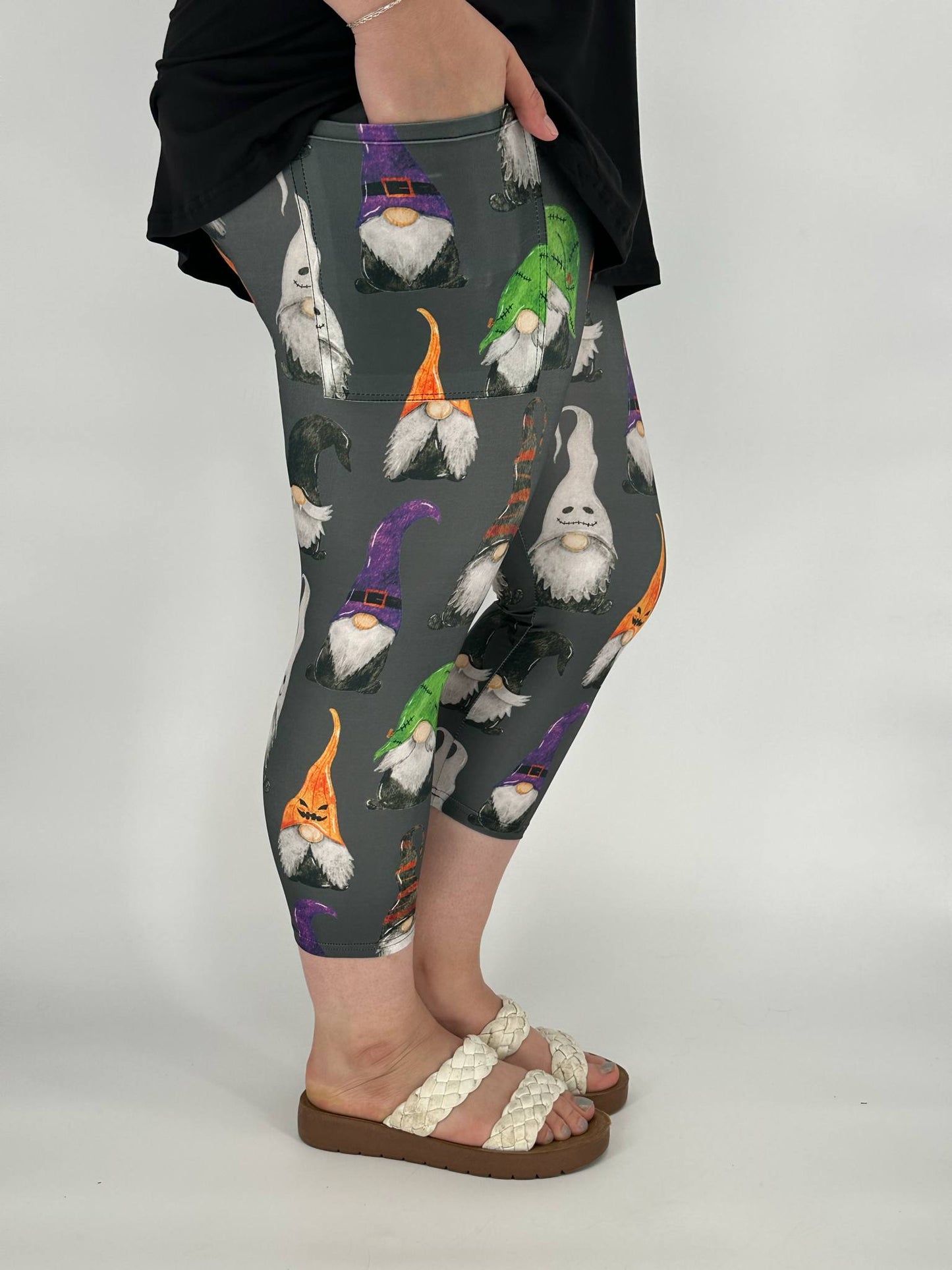 Model wearing gray capri leggings with Halloween gnome print featuring witch, ghost, and jack-o-lantern gnomes, with square side pockets, styled with a black top and cream sandals
