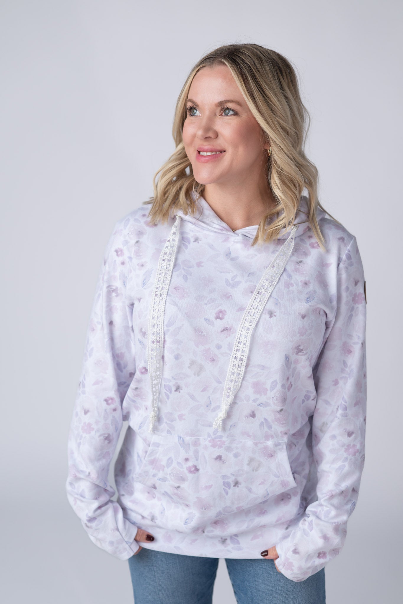 Woman wearing a white Hailey Pullover Hoodie with soft watercolor Blush Floral print in lavender and dusty pink, featuring crochet drawstrings and kangaroo pocket