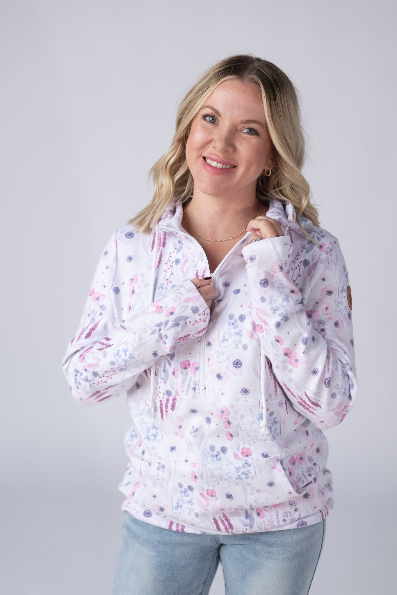 Woman wearing a white Half Zip Hoodie with soft watercolor Wildflower Gardens print in pink, purple, and blue, featuring a kangaroo pocket and hood with drawstrings