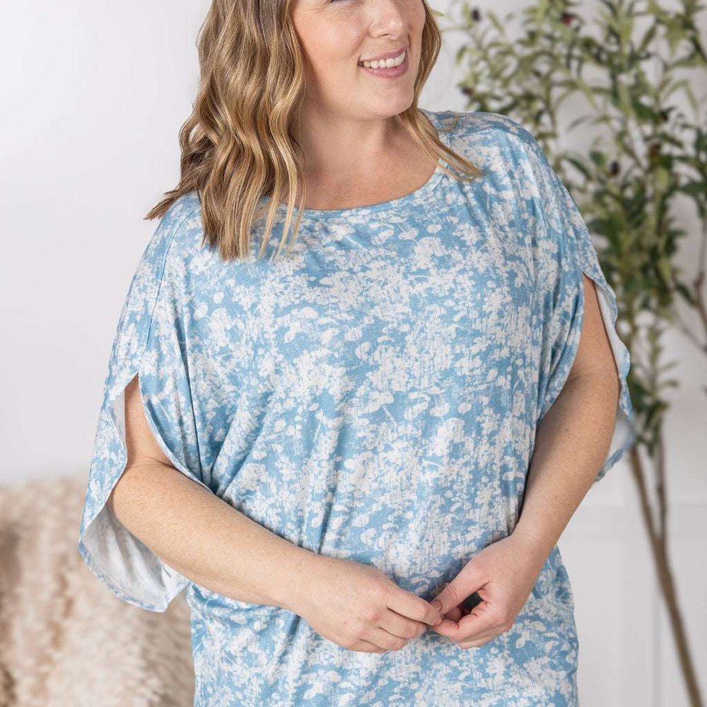 
                  
                    Close-up of a smiling model wearing the Darcy Dolman Top in a soft blue and ivory abstract print, featuring relaxed dolman sleeves and a rounded neckline.
                  
                