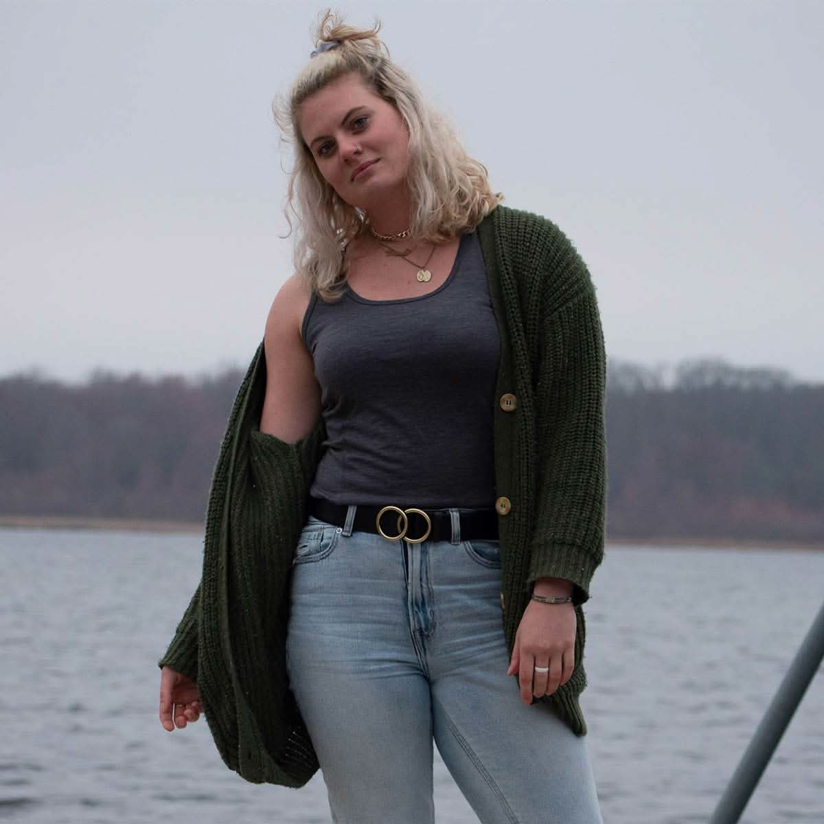 Woman standing by a body of water wearing a green cardigan, gray tank top, and light blue jeans.