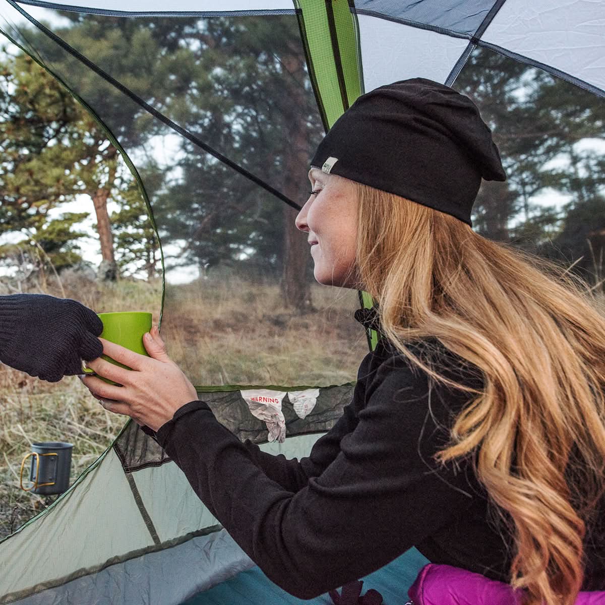 Woman in a black beanie and jacket holding a green mug, standing inside a tent with trees in the background.