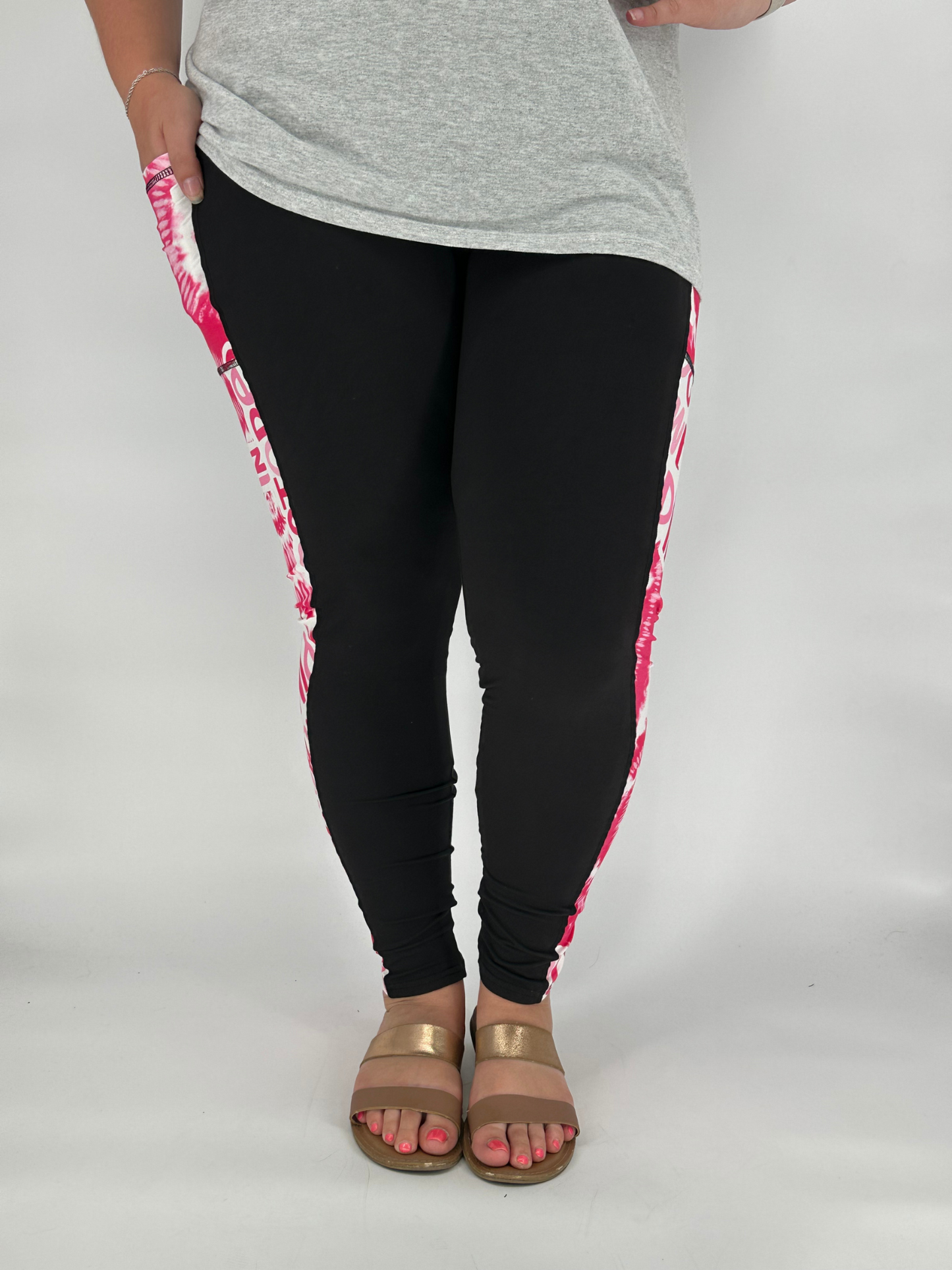 Model wearing black full-length leggings with pink and white tie-dye side panel featuring breast cancer awareness text, with square side pockets, styled with a grey top and gold sandals
