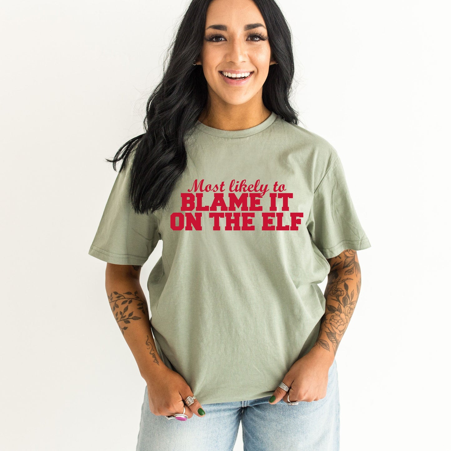 Model wearing sage green graphic tee with red text reading "Most Likely to Blame It on the Elf" styled with light wash jeans