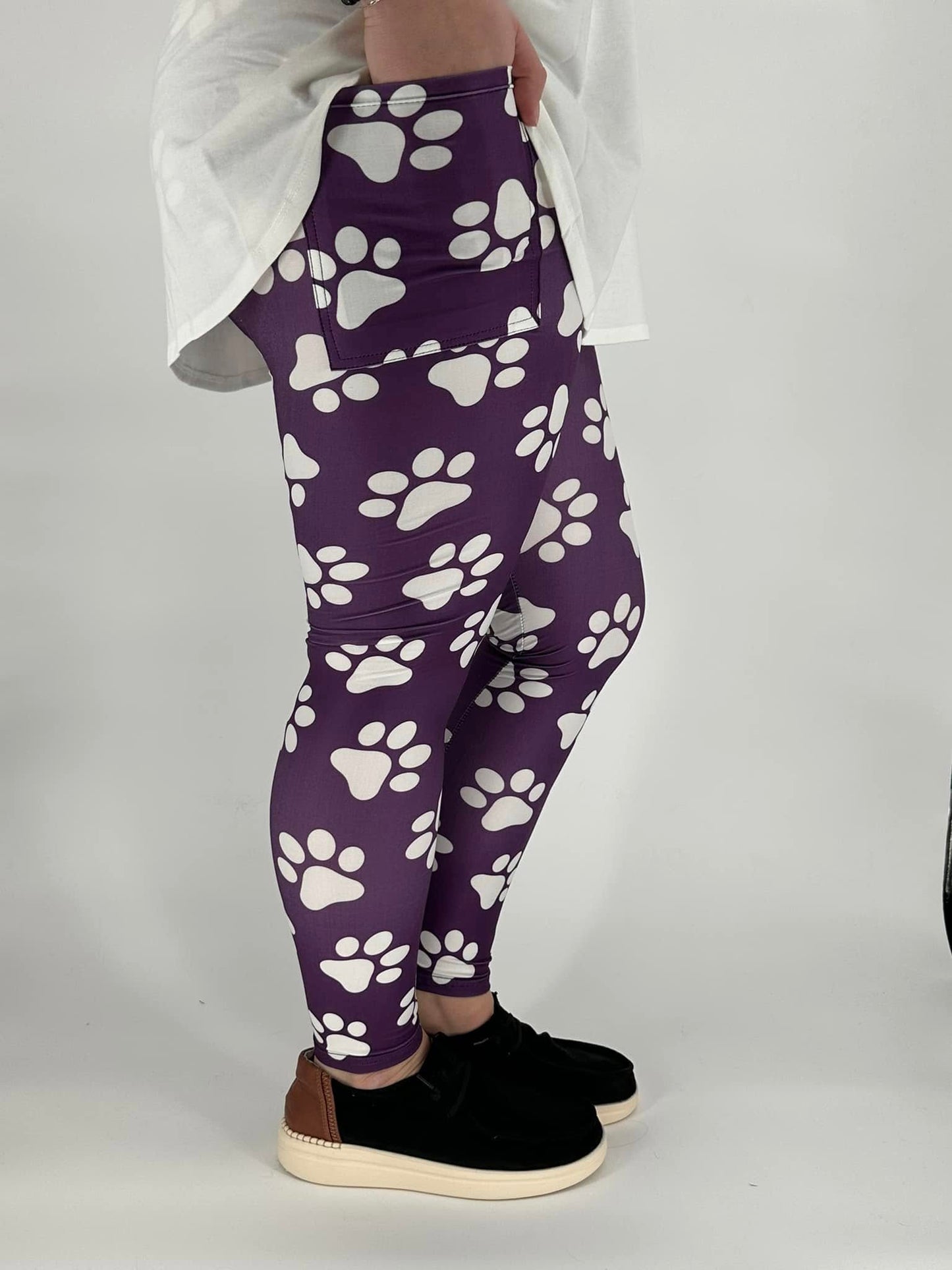 Model wearing plum purple full-length leggings with white paw print design and square side pockets, styled with a white long-sleeve top and black sneakers