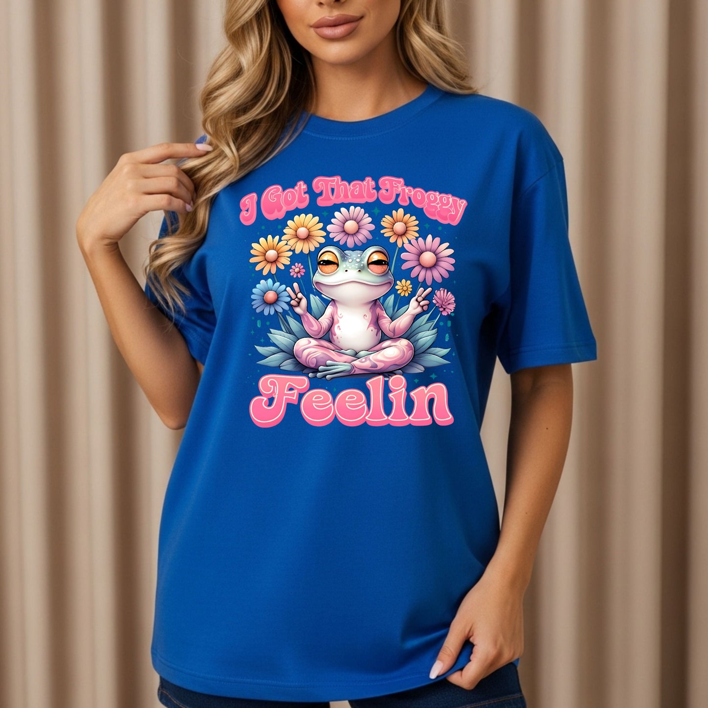 Royal blue graphic t-shirt featuring a pink frog in meditation pose with peace signs, surrounded by colorful daisies, with pink retro text reading "I Got That Froggy Feelin"