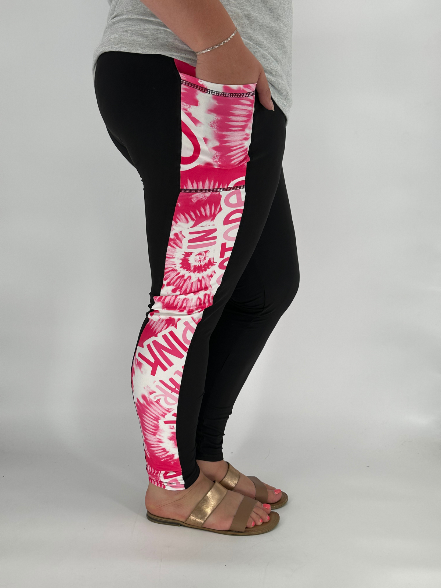 Model wearing black full-length leggings with pink and white tie-dye side panel featuring breast cancer awareness text, with square side pockets, styled with a grey top and gold sandals