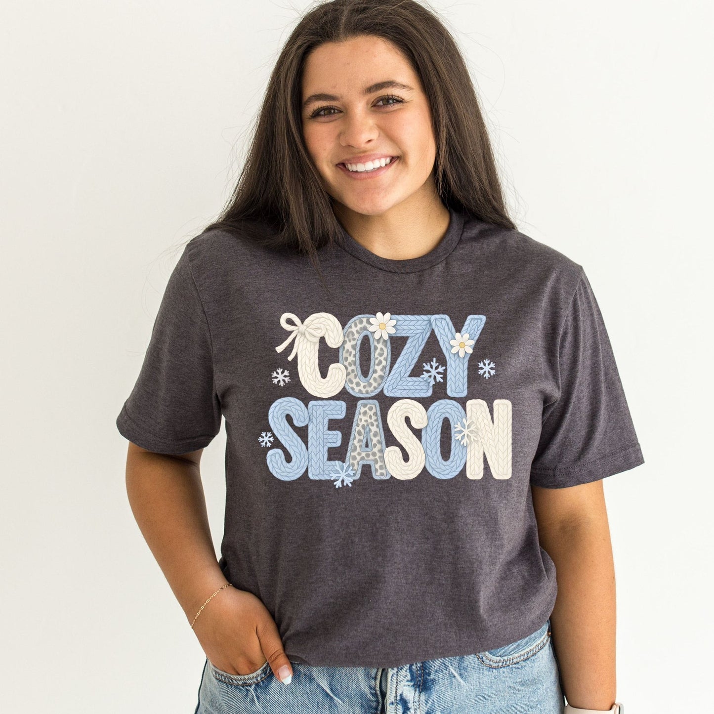 Model wearing charcoal heather graphic tee with "Cozy Season" in knit-textured lettering in soft blue, cream, and leopard print with snowflakes and daisies, styled with light wash jeans