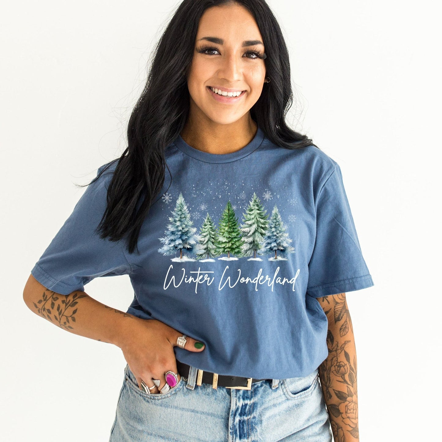 Model wearing indigo blue graphic tee with snowy pine trees and "Winter Wonderland" script in white, styled with light wash jeans and black belt