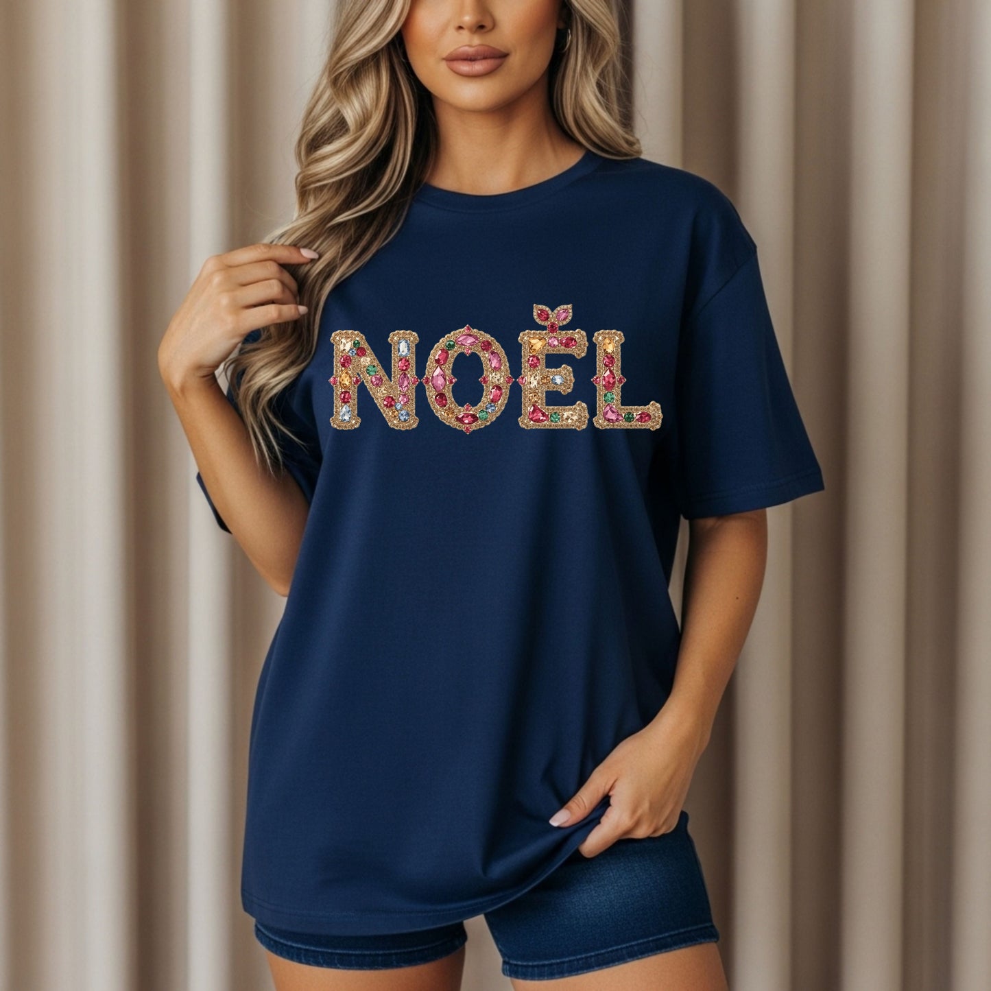 Model wearing navy blue graphic tee with "NOËL" spelled in sparkling jeweled letters featuring gold, pink, red, and multicolored gems