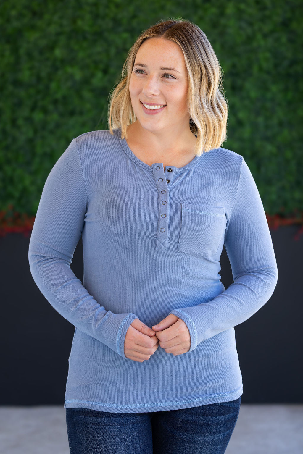 Woman wearing a blue long-sleeve shirt with a pocket against a green wall.