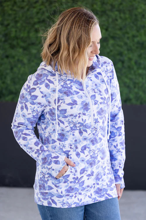 Woman wearing half-zip hoodie with blue watercolor floral print all over, featuring kangaroo pocket, drawstring hood, and half-zip closure, paired with jeans