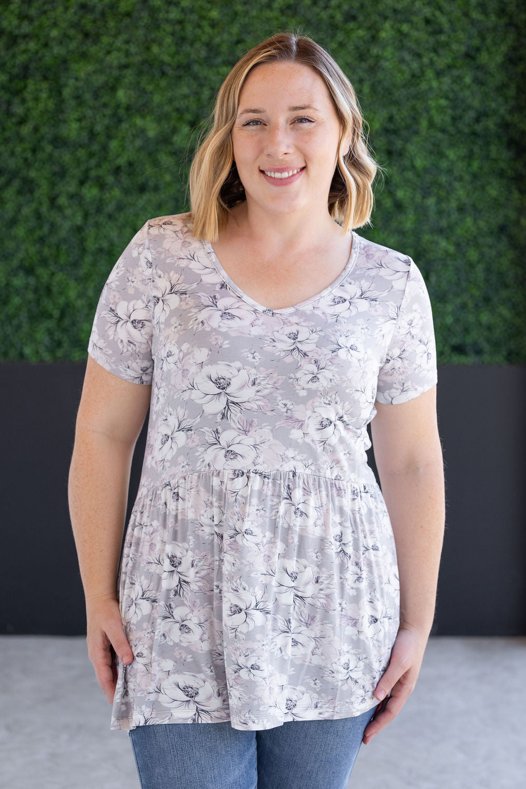 Woman wearing short sleeve ruffle top with vintage grey and white floral print and gathered ruffle hem at waist, paired with dark jeans