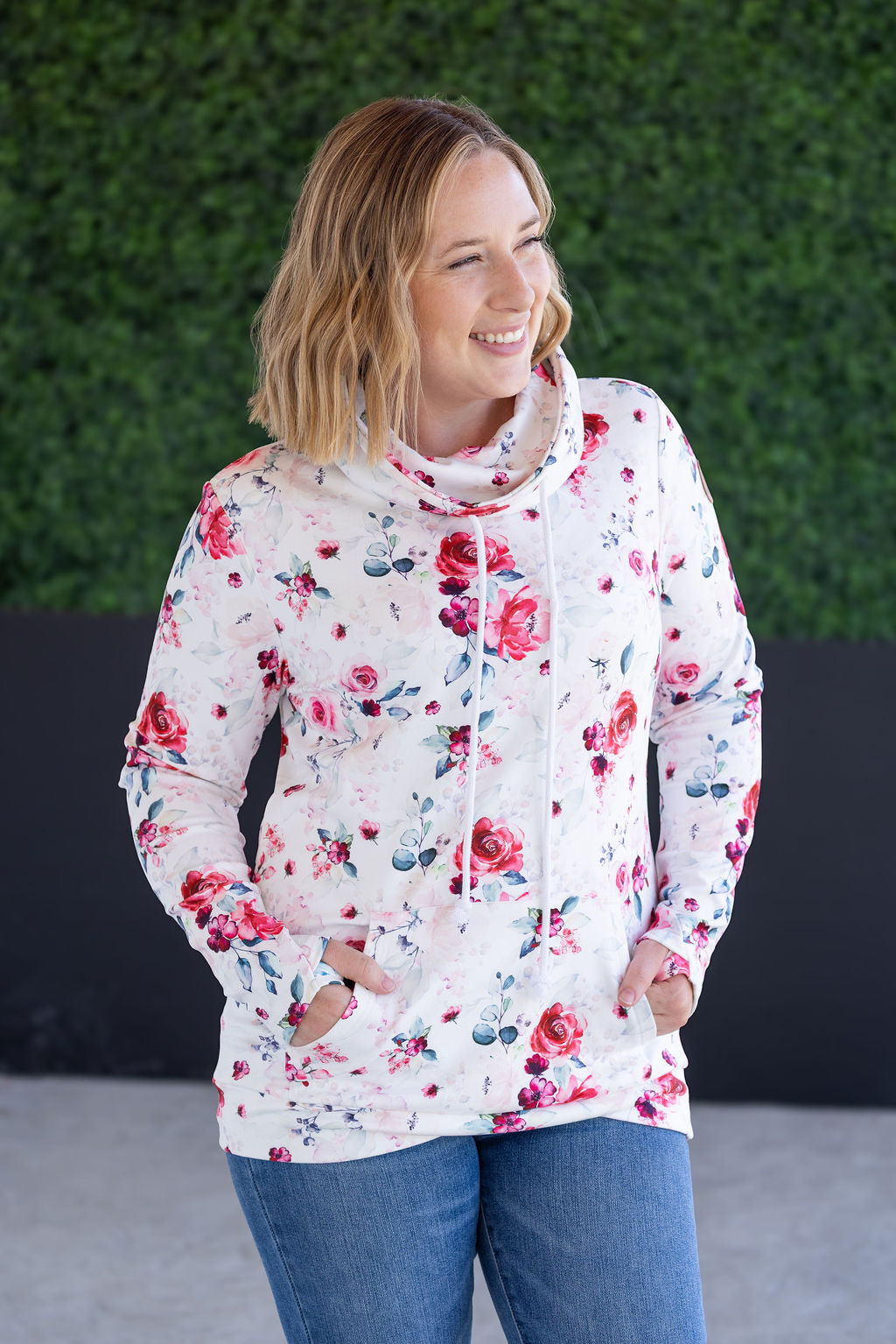 Woman wearing white funnel neck sweatshirt with pink floral print, drawstring neckline, and kangaroo pocket