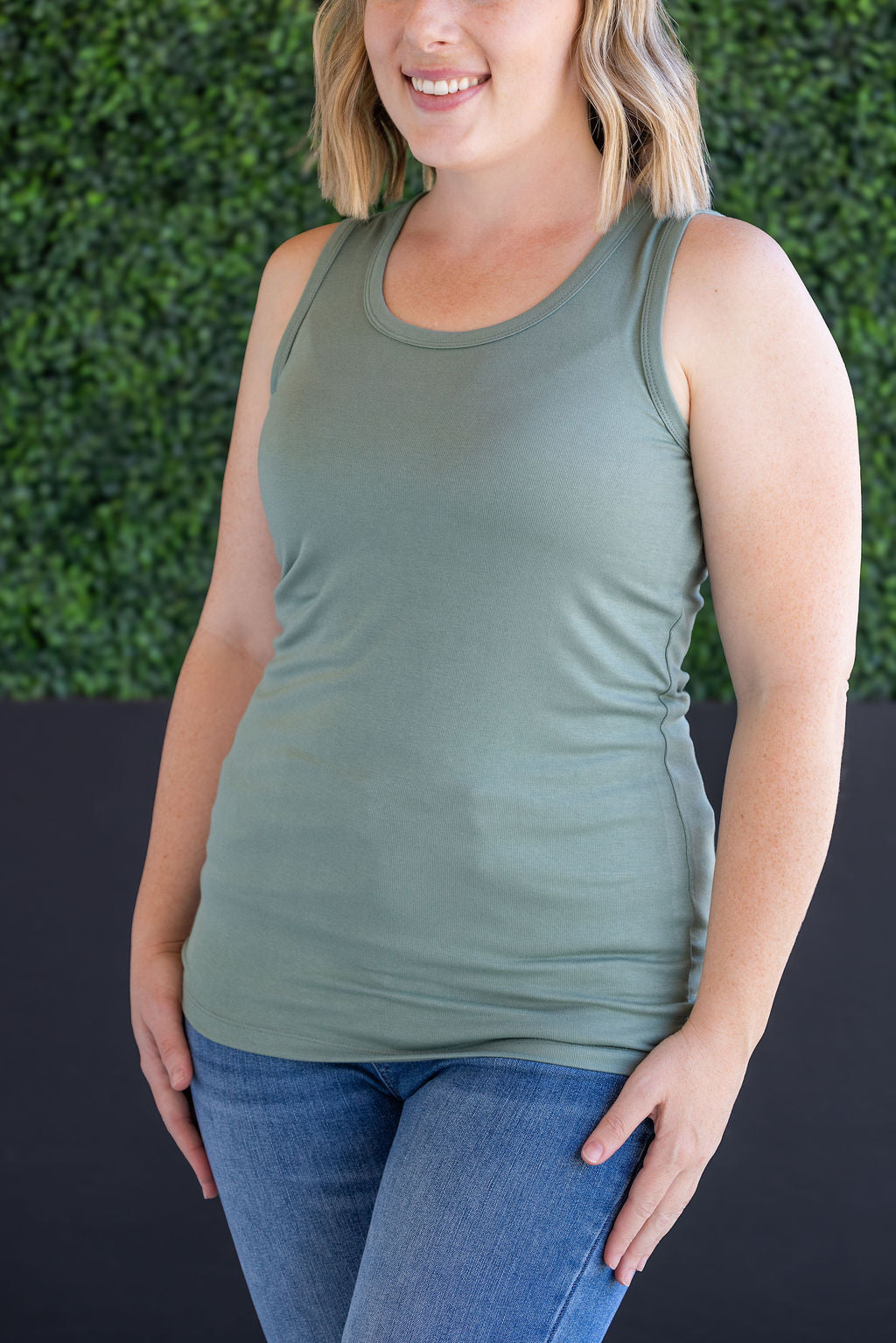 Woman wearing sage green ribbed tank top with scoop neckline and fitted silhouette, paired with blue jeans