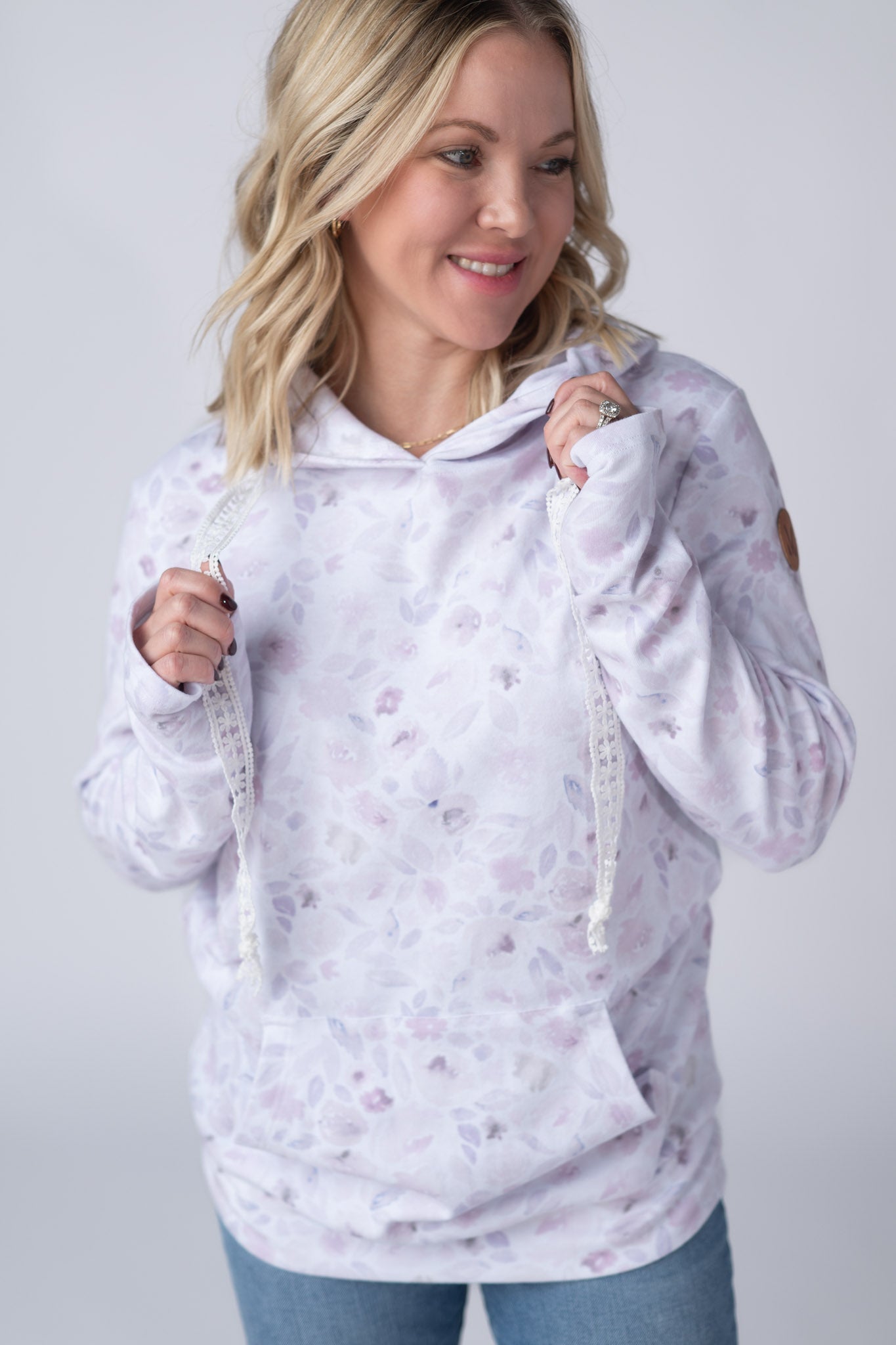 Woman wearing a white Hailey Pullover Hoodie with soft watercolor Blush Floral print in lavender and dusty pink, featuring crochet drawstrings and kangaroo pocket