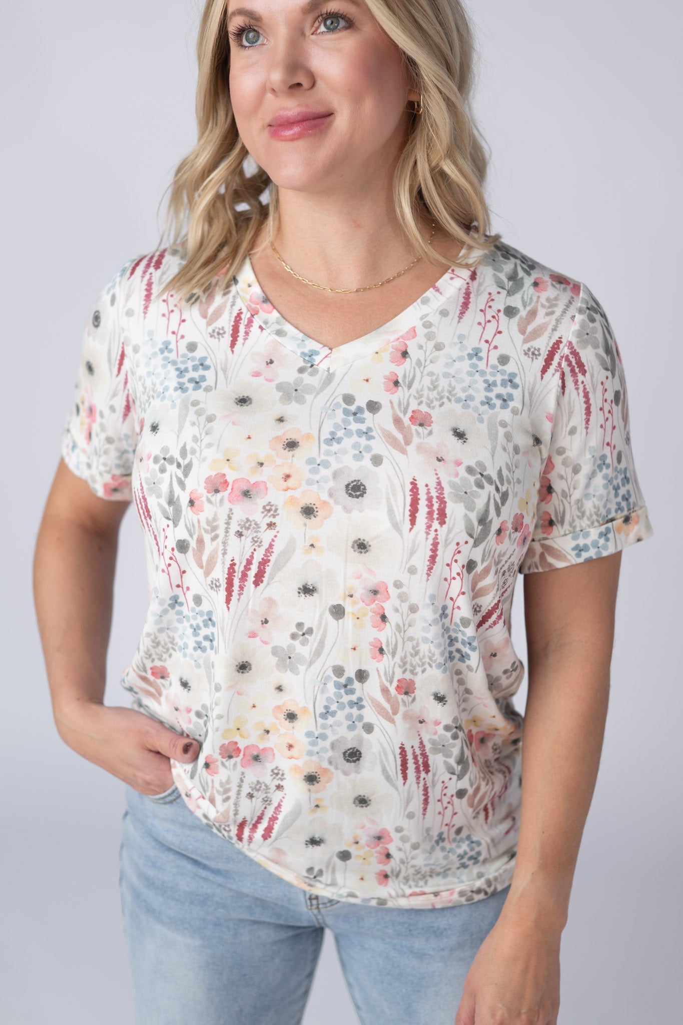 Woman wearing a white Sophie Tee with soft watercolor wildflower print in pink, peach, gray, and blue, with v-neckline and cuffed sleeves, paired with jeans