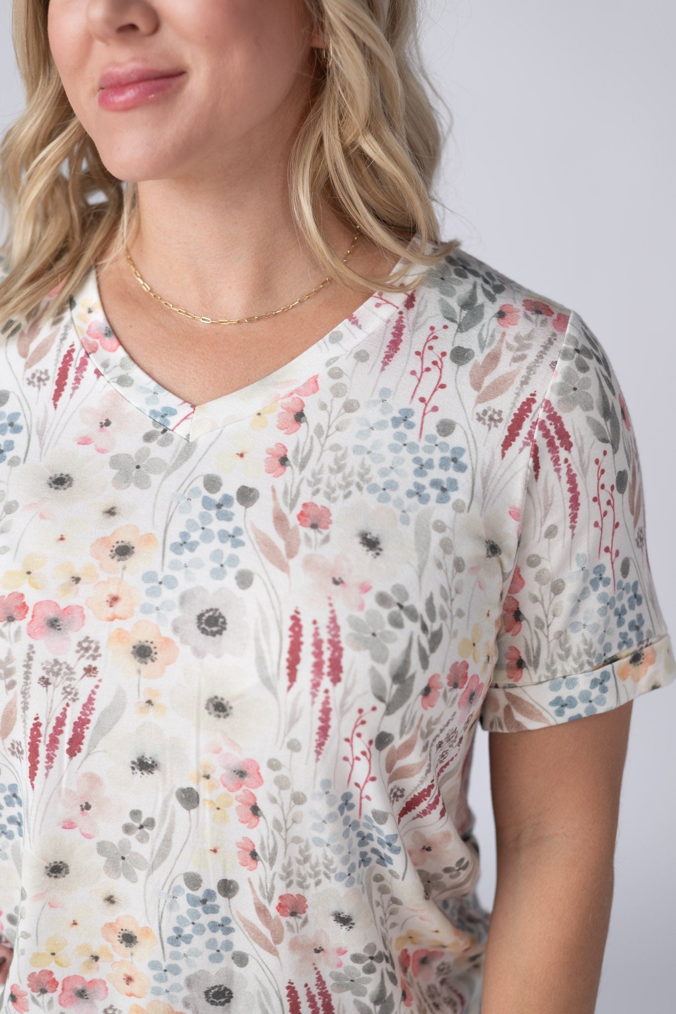 Woman wearing a white Sophie Tee with soft watercolor wildflower print in pink, peach, gray, and blue, with v-neckline and cuffed sleeves, paired with jeans