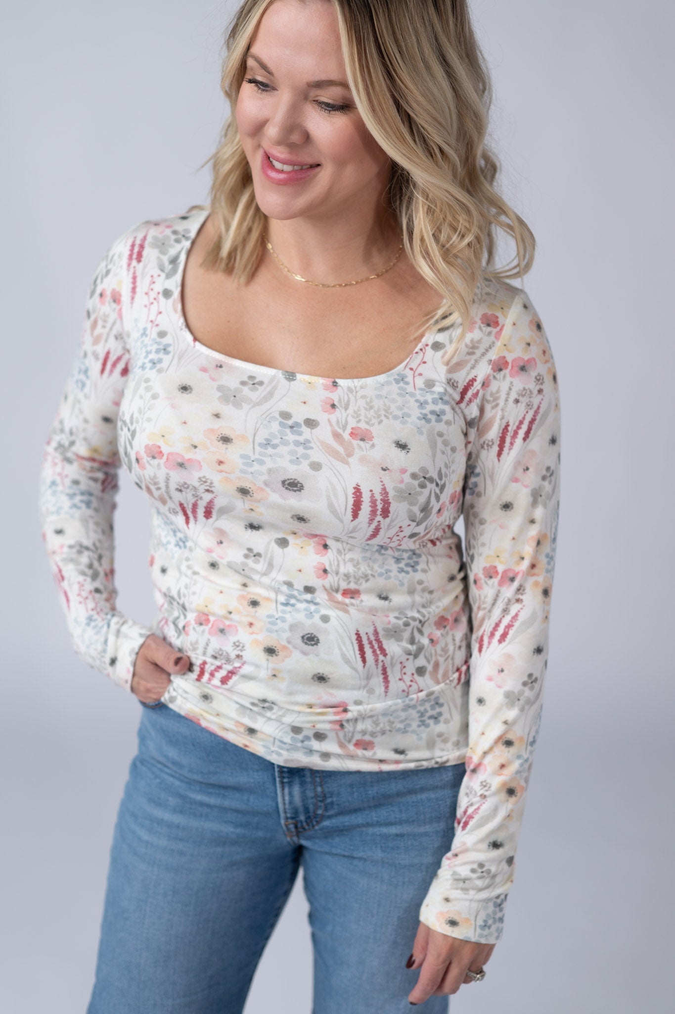 Woman wearing a white Alyssa Long Sleeve Top with soft watercolor wildflower print in pink, peach, gray, and blue, featuring a square neckline, paired with jeans