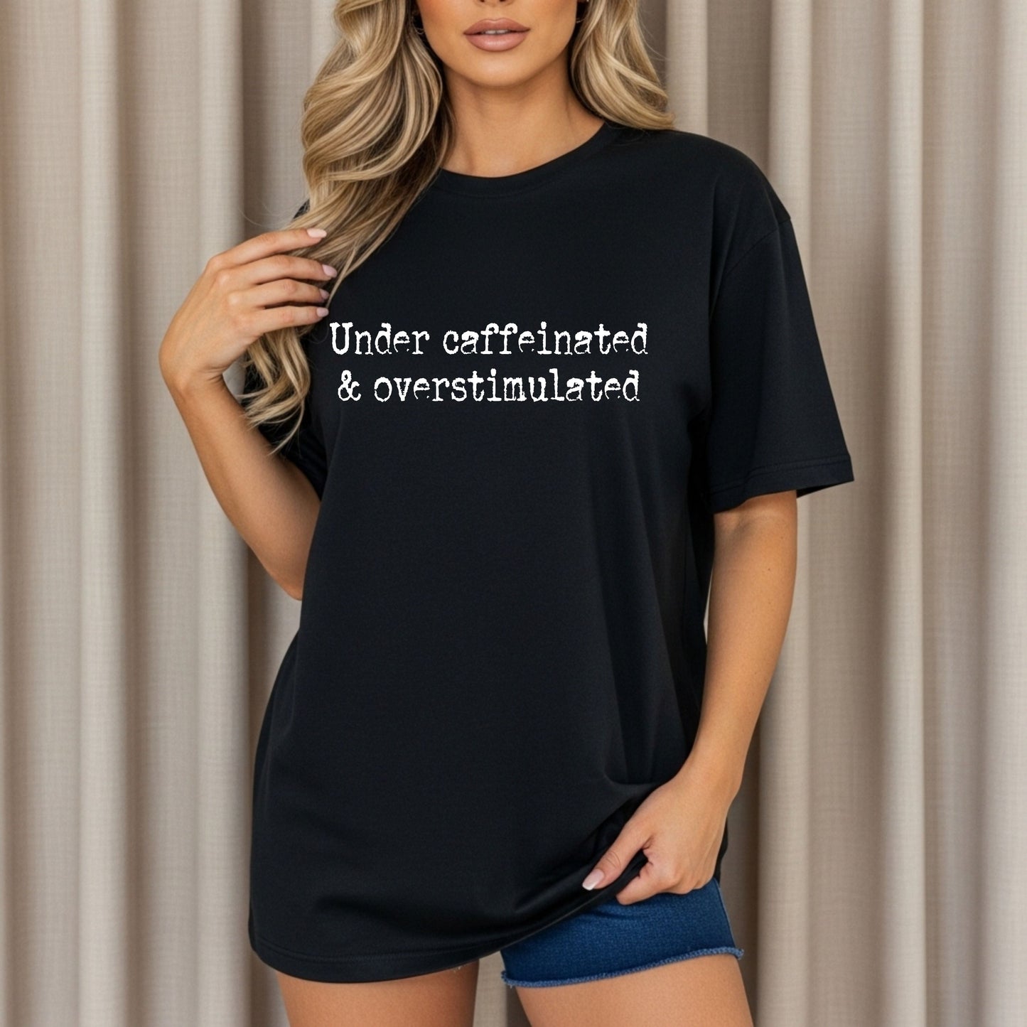 Person wearing a black t-shirt with text 'Under caffeinated & overstimulated' against a beige curtain background