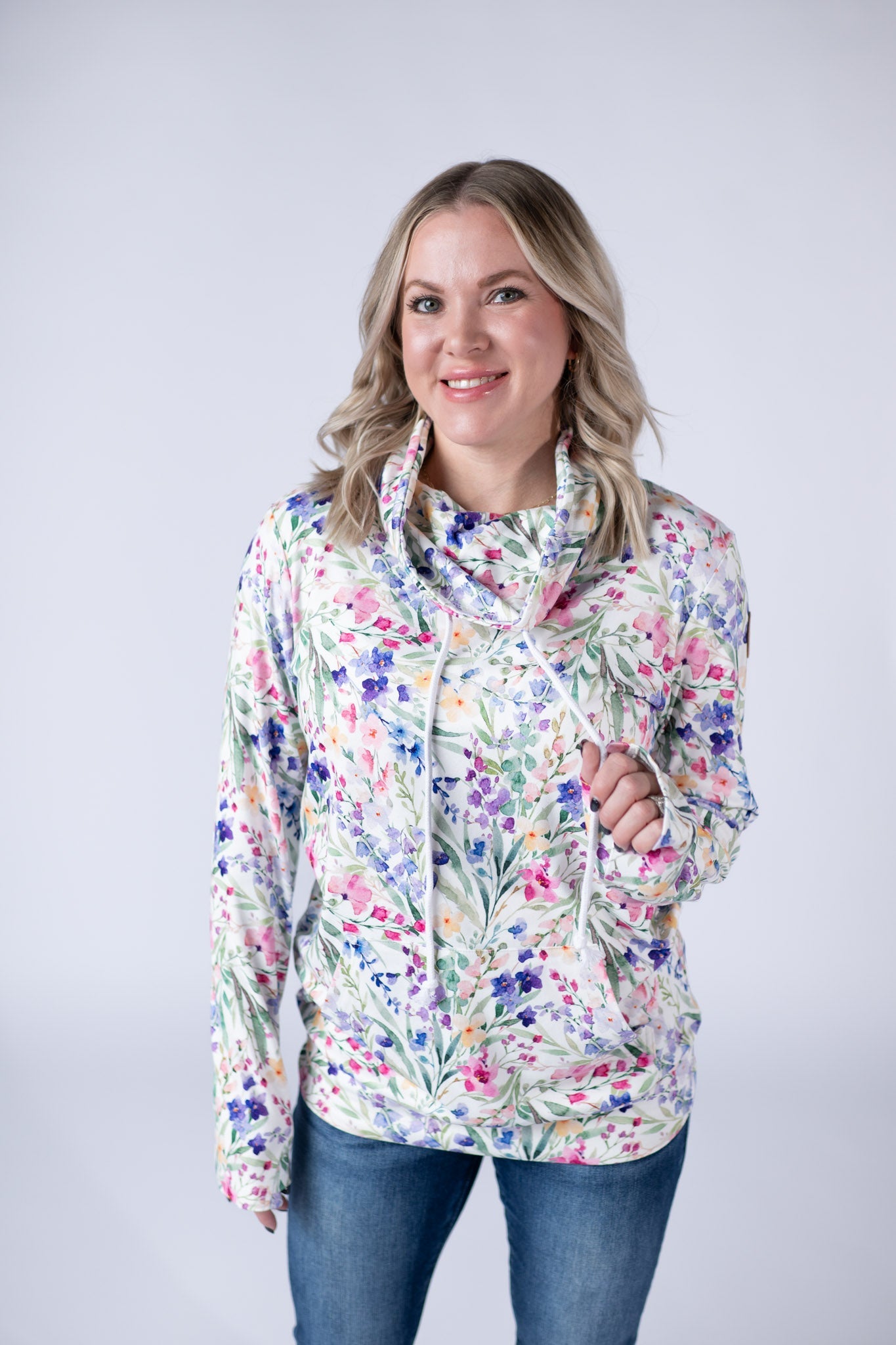 Woman wearing white funnel neck sweatshirt with pink, purple, and blue watercolor wildflower print and kangaroo pocket