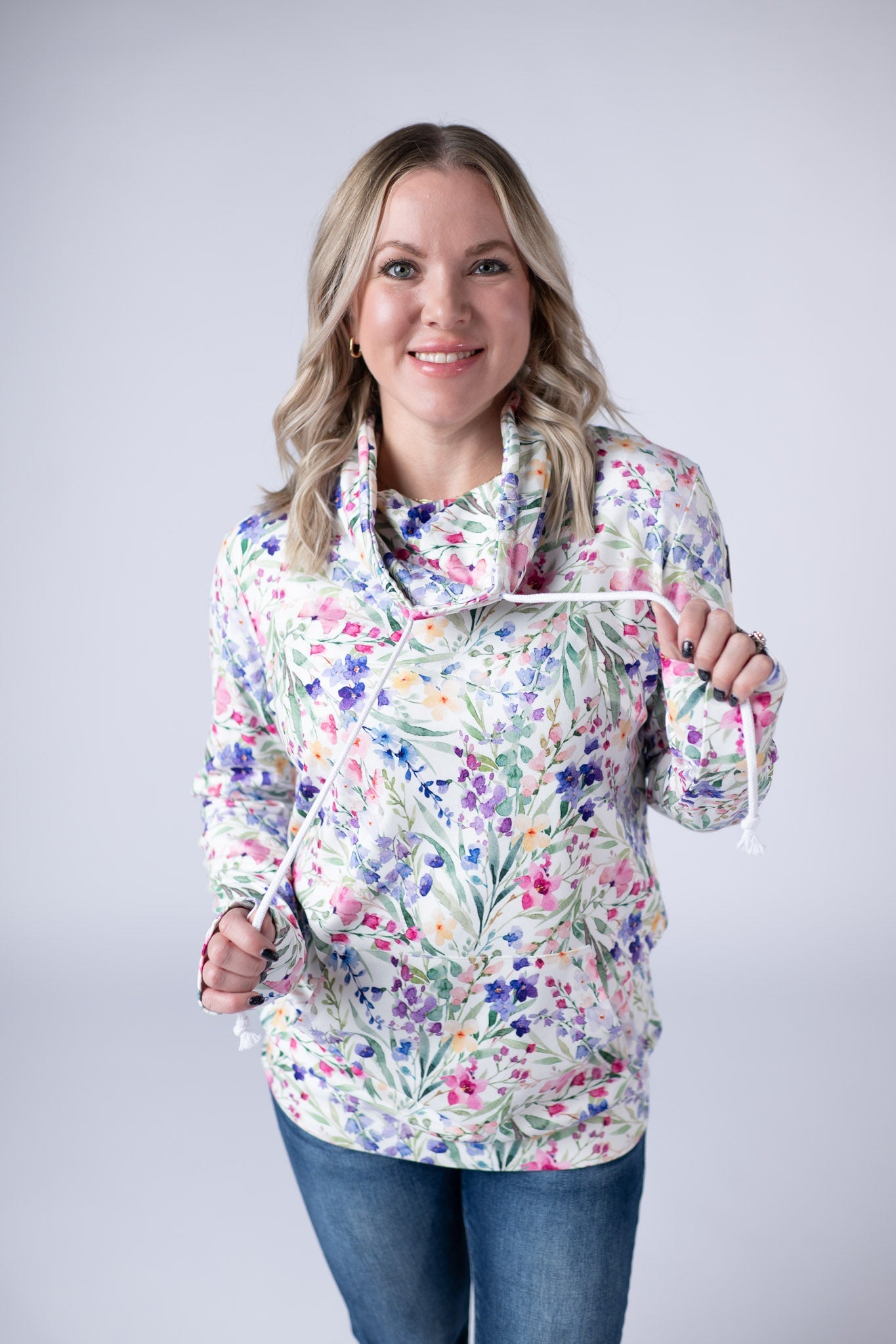 Woman wearing white funnel neck sweatshirt with pink, purple, and blue watercolor wildflower print and kangaroo pocket