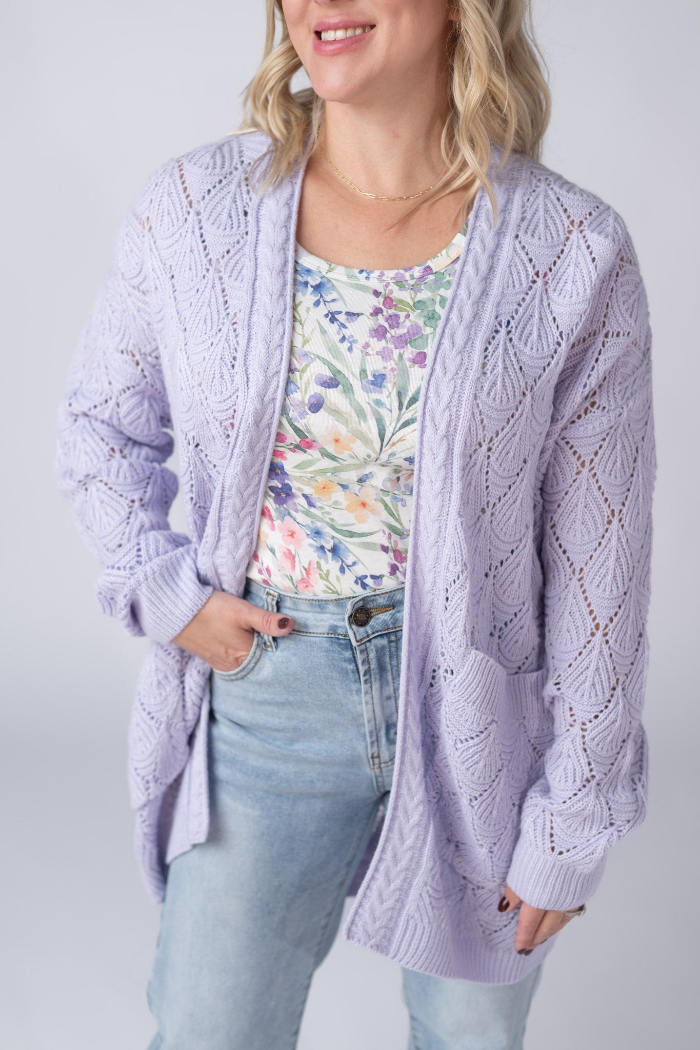 Woman wearing a lavender Open Knit Cardigan with leaf pattern detail and pockets, layered over a floral tank and jeans