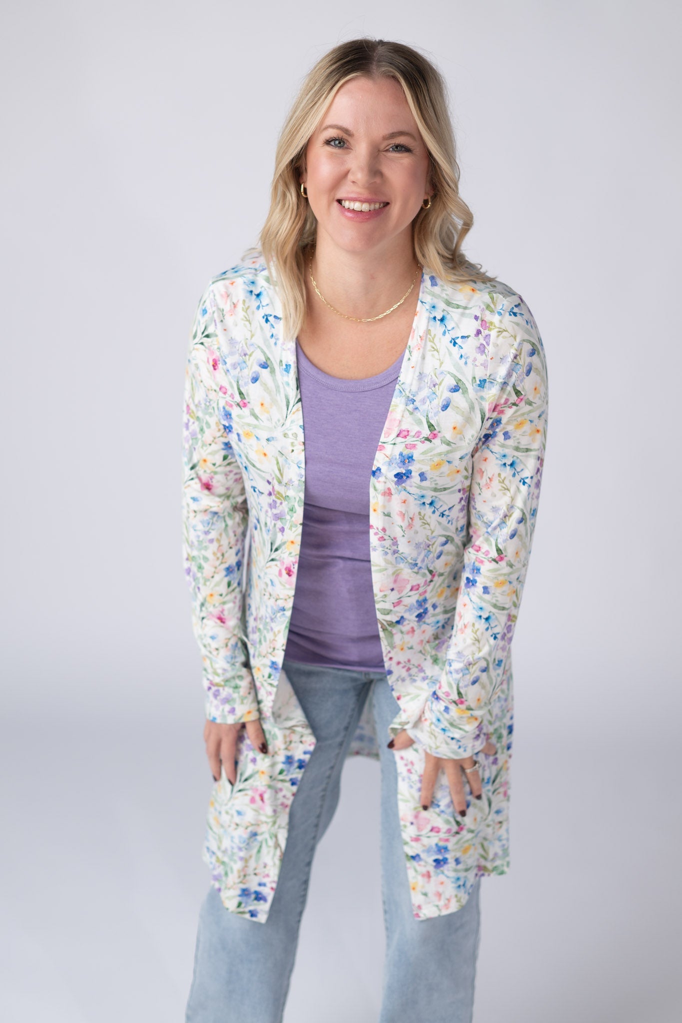 Woman wearing a white Classic Cardigan with bright watercolor cottage floral print in blue, purple, pink, yellow, and green, featuring an open front and pockets, styled over a lavender top with jeans