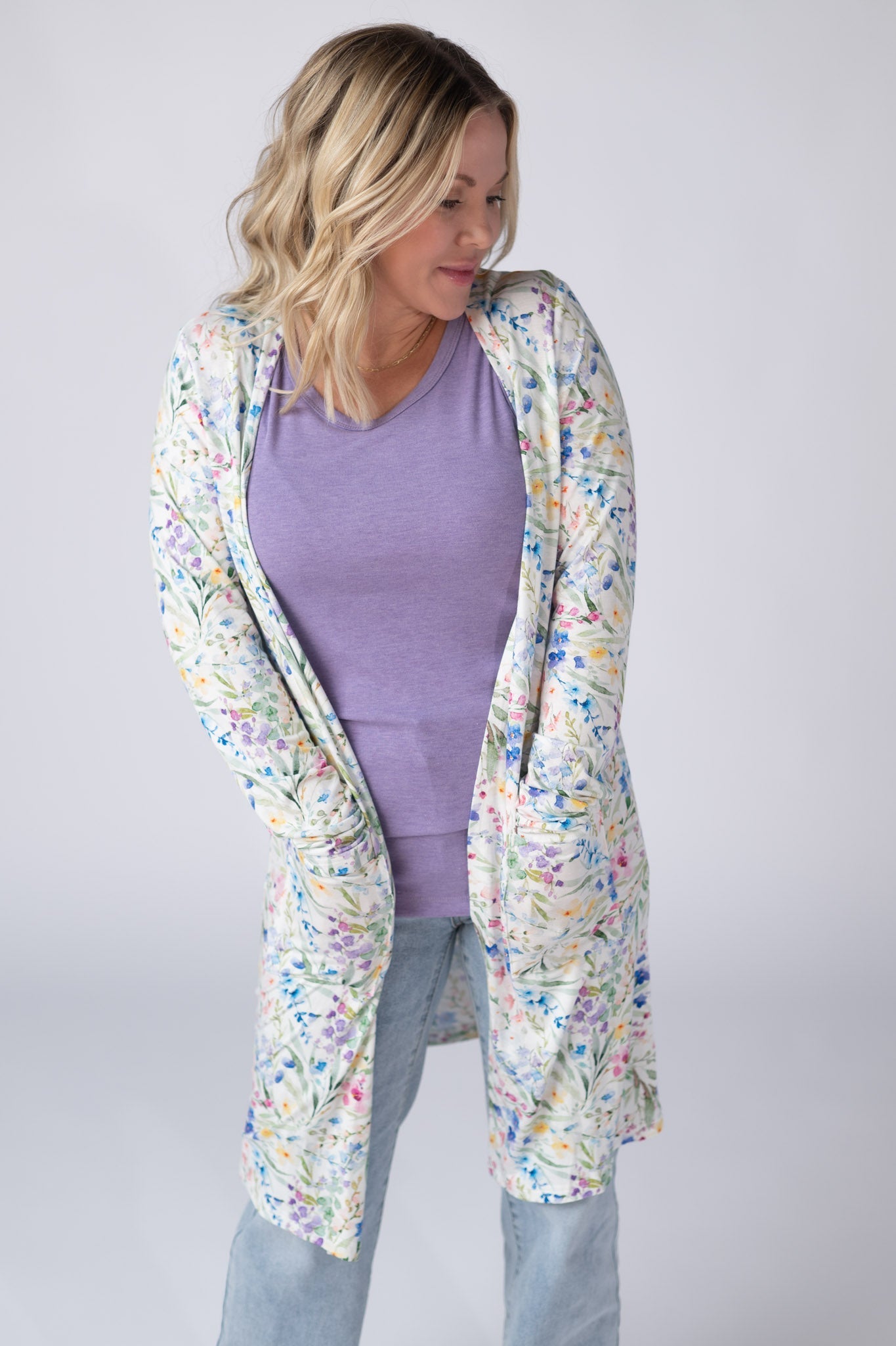 Woman wearing a white Classic Cardigan with bright watercolor cottage floral print in blue, purple, pink, yellow, and green, featuring an open front and pockets, styled over a lavender top with jeans