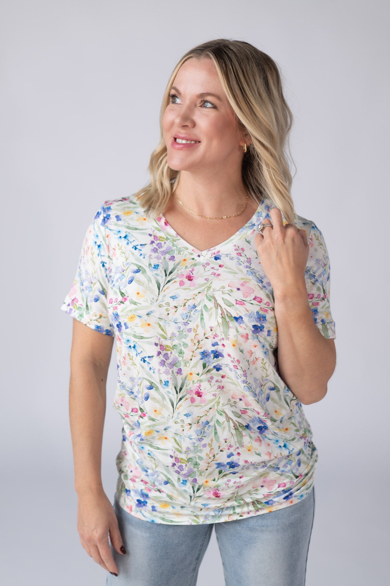 Woman wearing a white Sophie Tee with bright watercolor cottage floral print in blue, purple, pink, yellow, and green, with v-neckline and cuffed sleeves, paired with jeans