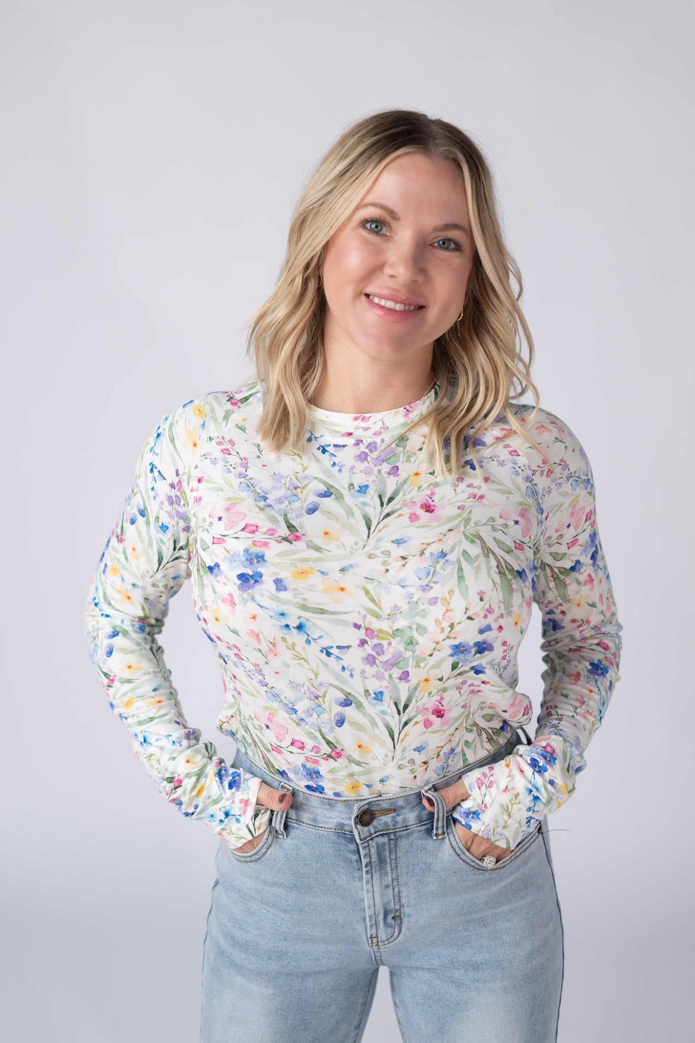 Woman wearing a white Blair Long Sleeve Top with bright watercolor cottage floral print in blue, purple, pink, yellow, and green, paired with jeans