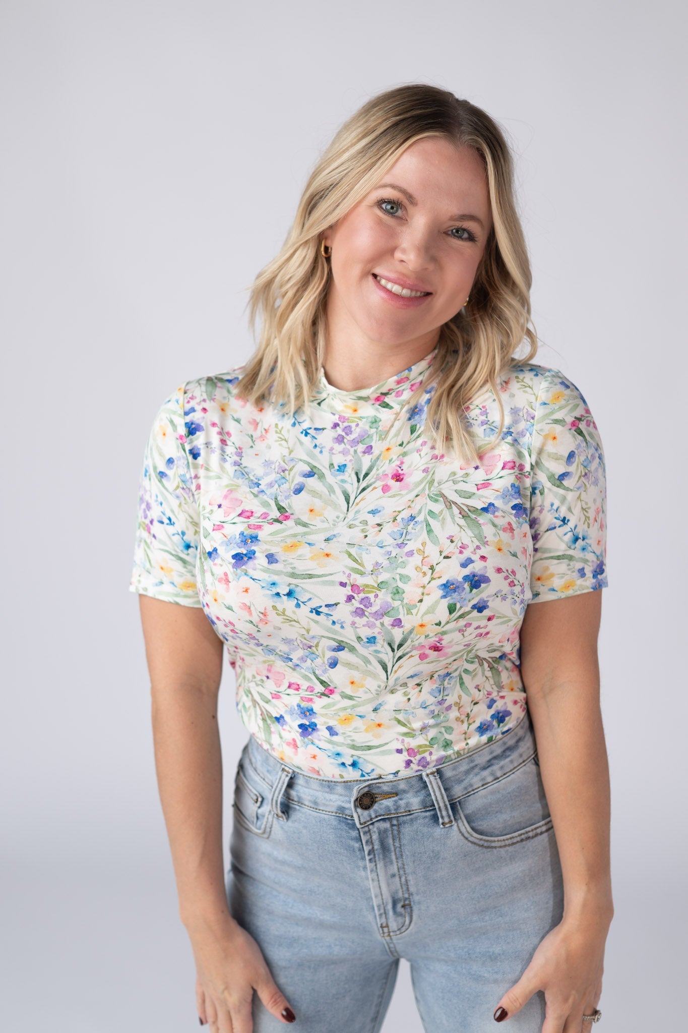 Woman wearing a white Blair Short Sleeve Top with bright watercolor cottage floral print in blue, purple, pink, yellow, and green, paired with jeans