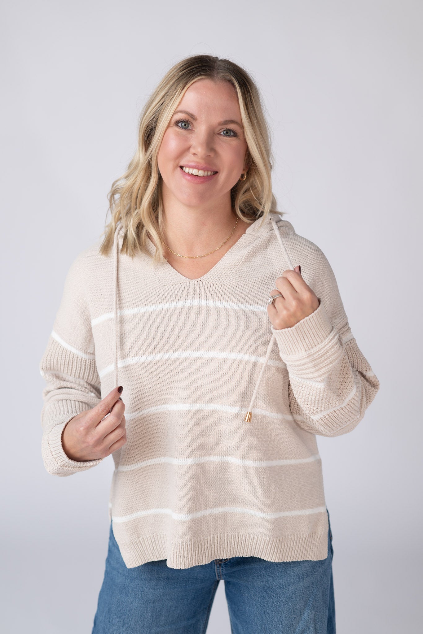 Woman wearing a natural beige Maddie Striped Hoodie with white stripes, v-neckline, hood with drawstrings, paired with jeans