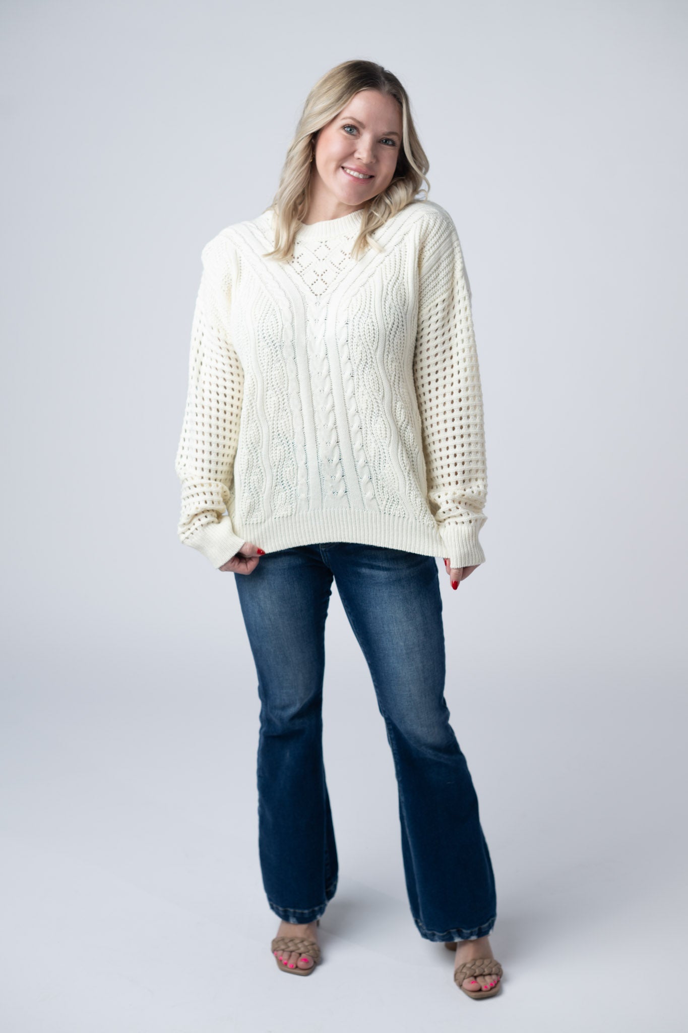 Woman wearing an ivory Jasmine Open Knit Sweater with cable knit details and open knit sleeves, paired with dark wash jeans