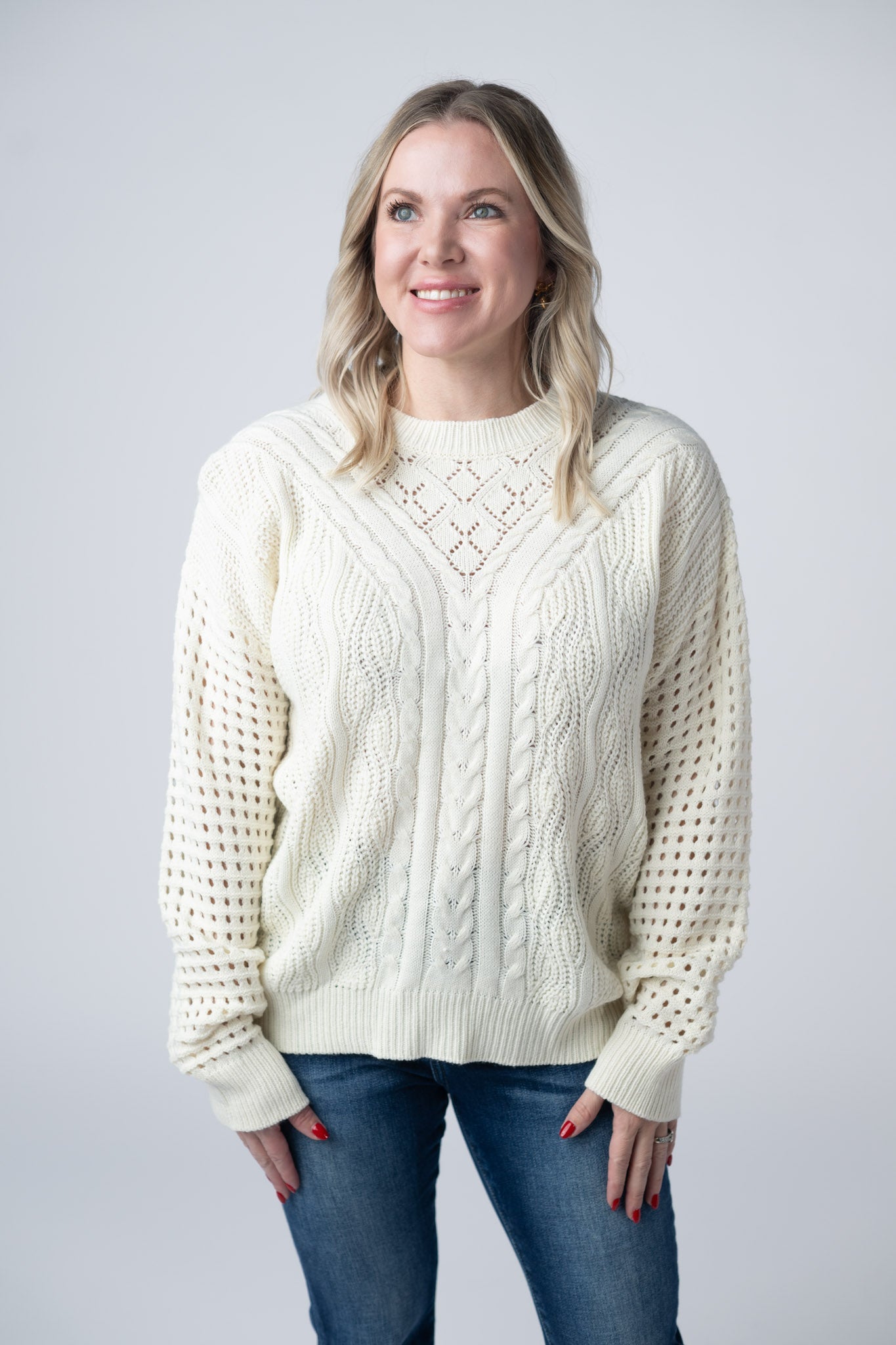 Woman wearing an ivory Jasmine Open Knit Sweater with cable knit details and open knit sleeves, paired with dark wash jeans