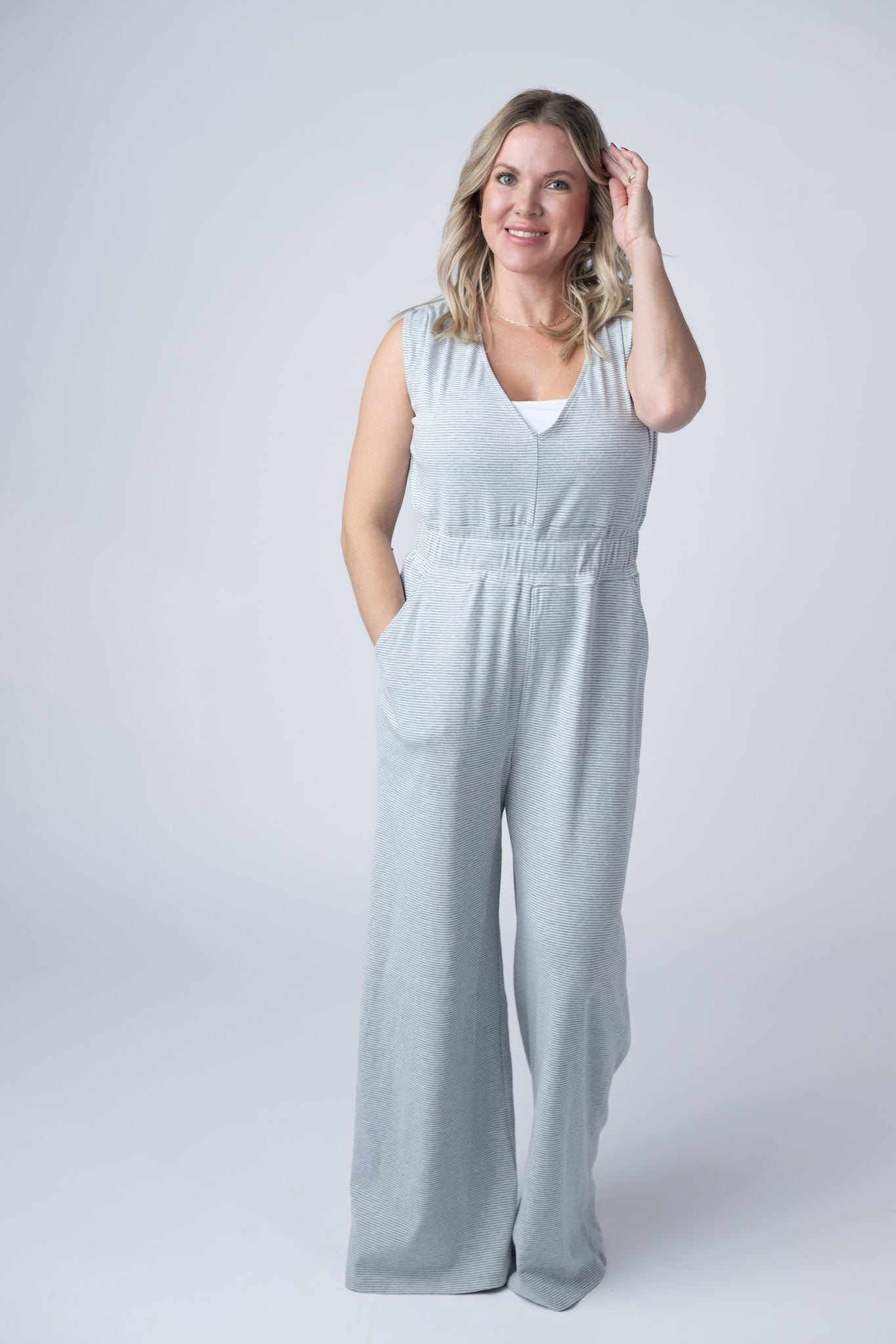 Women's wide leg jumpsuit in gray and white stripes with V-neckline, ruched shoulders, elastic waist, and side pockets styled with sandals