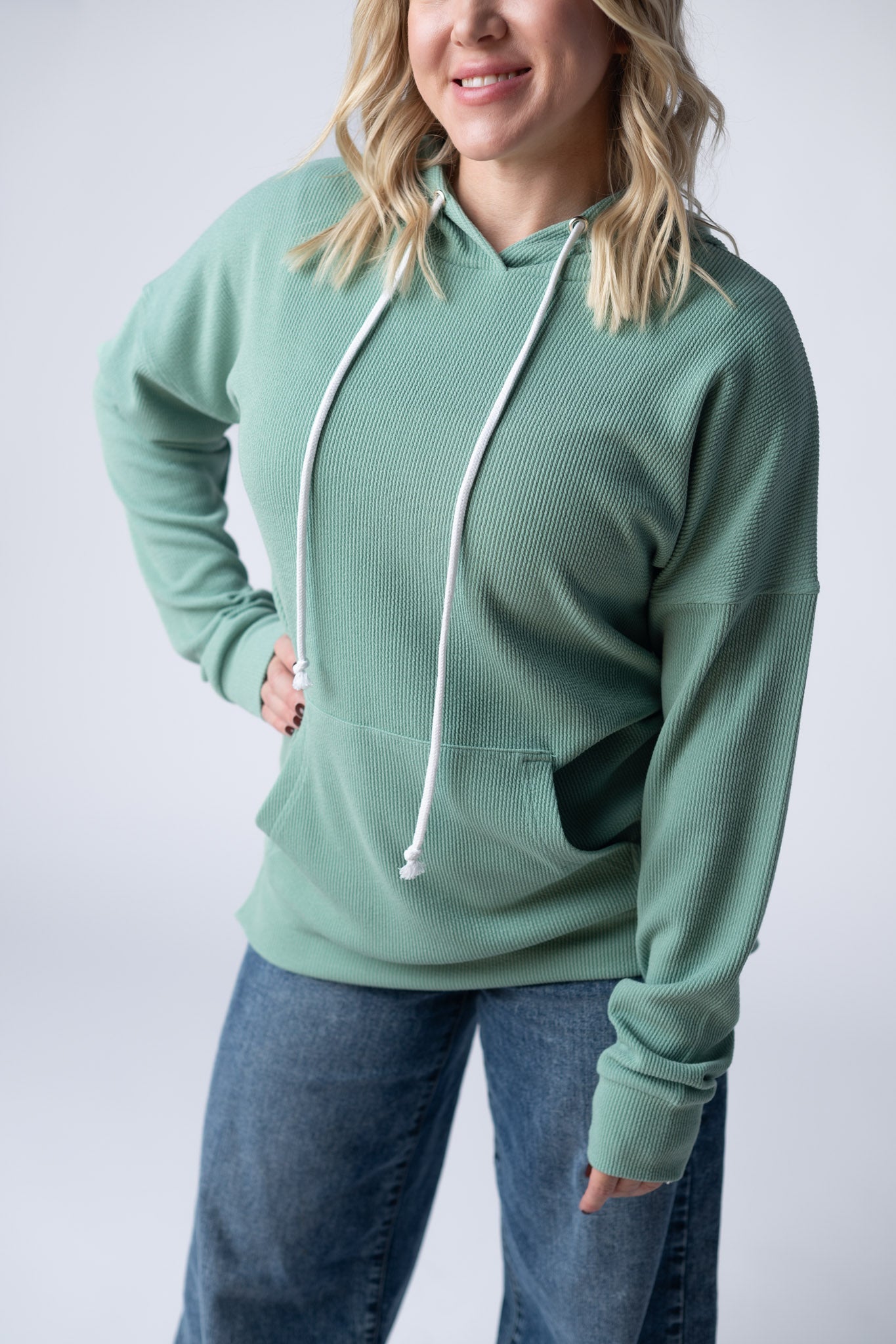 Woman wearing a sage green Kacey Ribbed Hoodie with white drawstrings, kangaroo pocket, and ribbed texture, paired with jeans