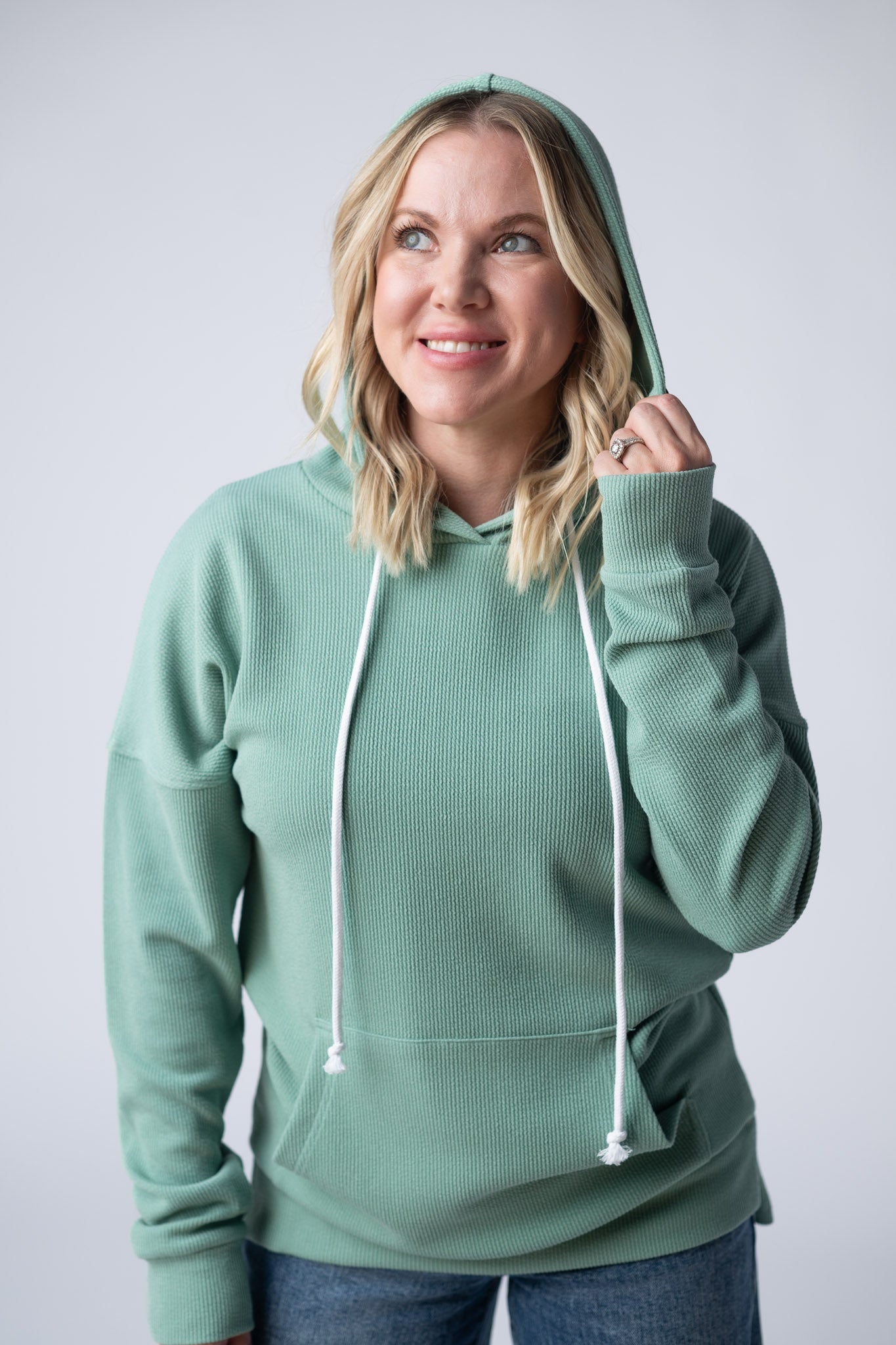 Woman wearing a sage green Kacey Ribbed Hoodie with white drawstrings, kangaroo pocket, and ribbed texture, paired with jeans