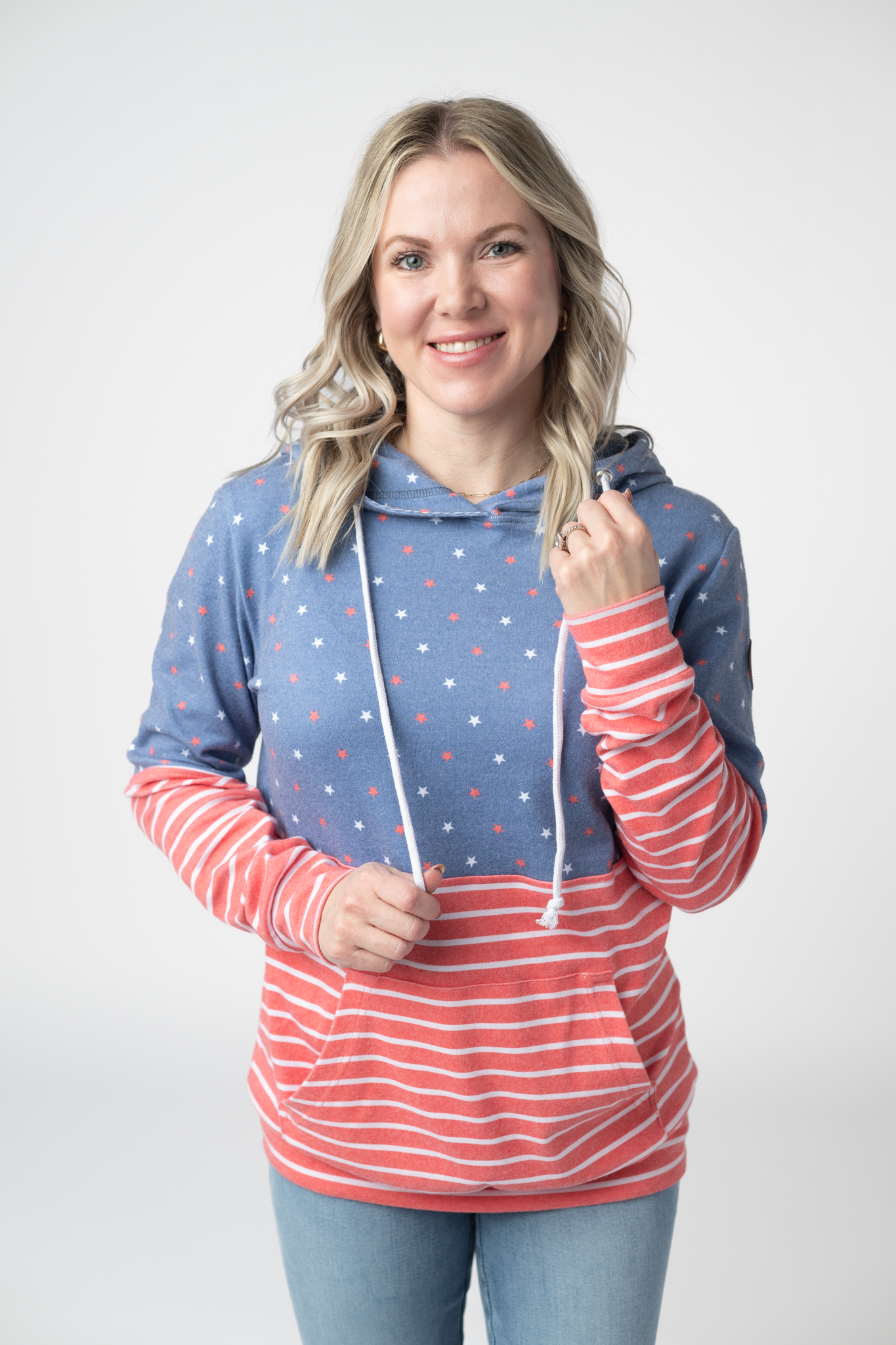 Woman wearing a colorblock pullover hoodie with faded blue star print on top and red and white stripes on the body and sleeves, featuring kangaroo pocket and white drawstrings
