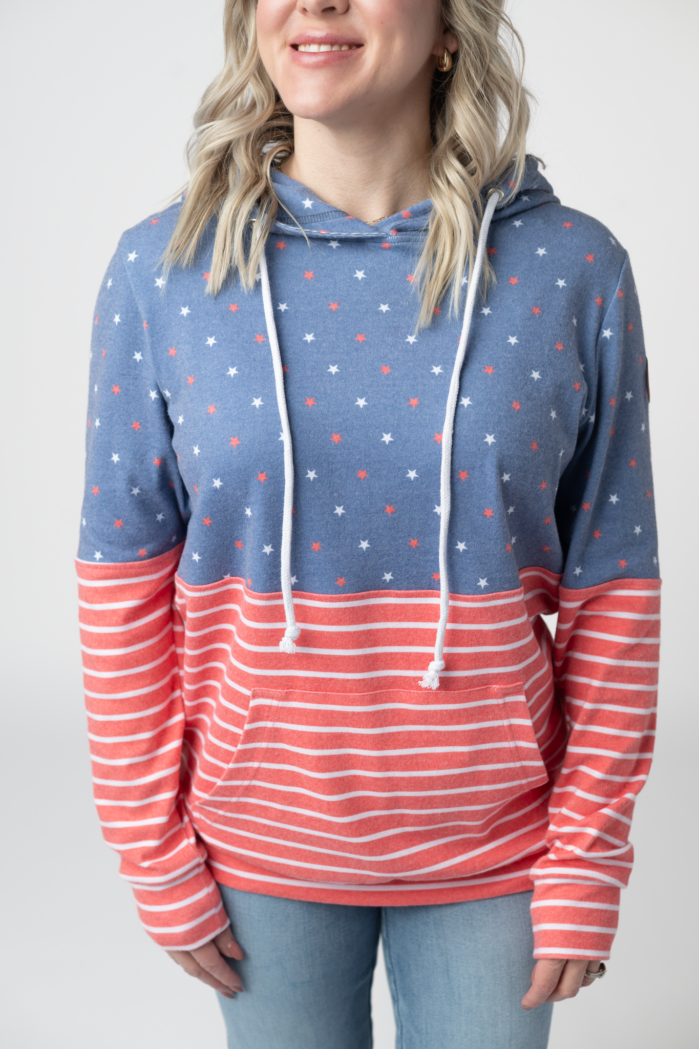 Woman wearing a colorblock pullover hoodie with faded blue star print on top and red and white stripes on the body and sleeves, featuring kangaroo pocket and white drawstrings