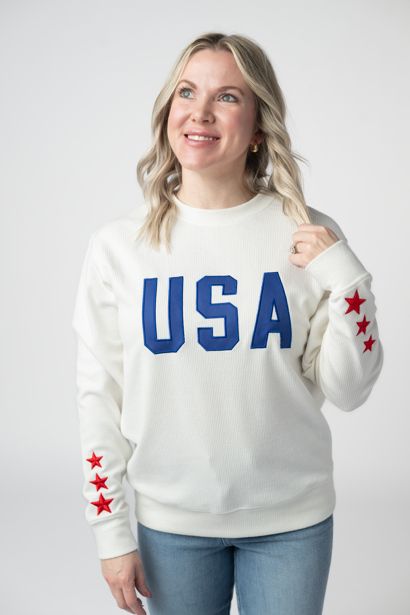 Woman wearing a white ribbed crew neck pullover sweatshirt with blue USA applique lettering on chest and red star accents on sleeves, paired with light wash jeans