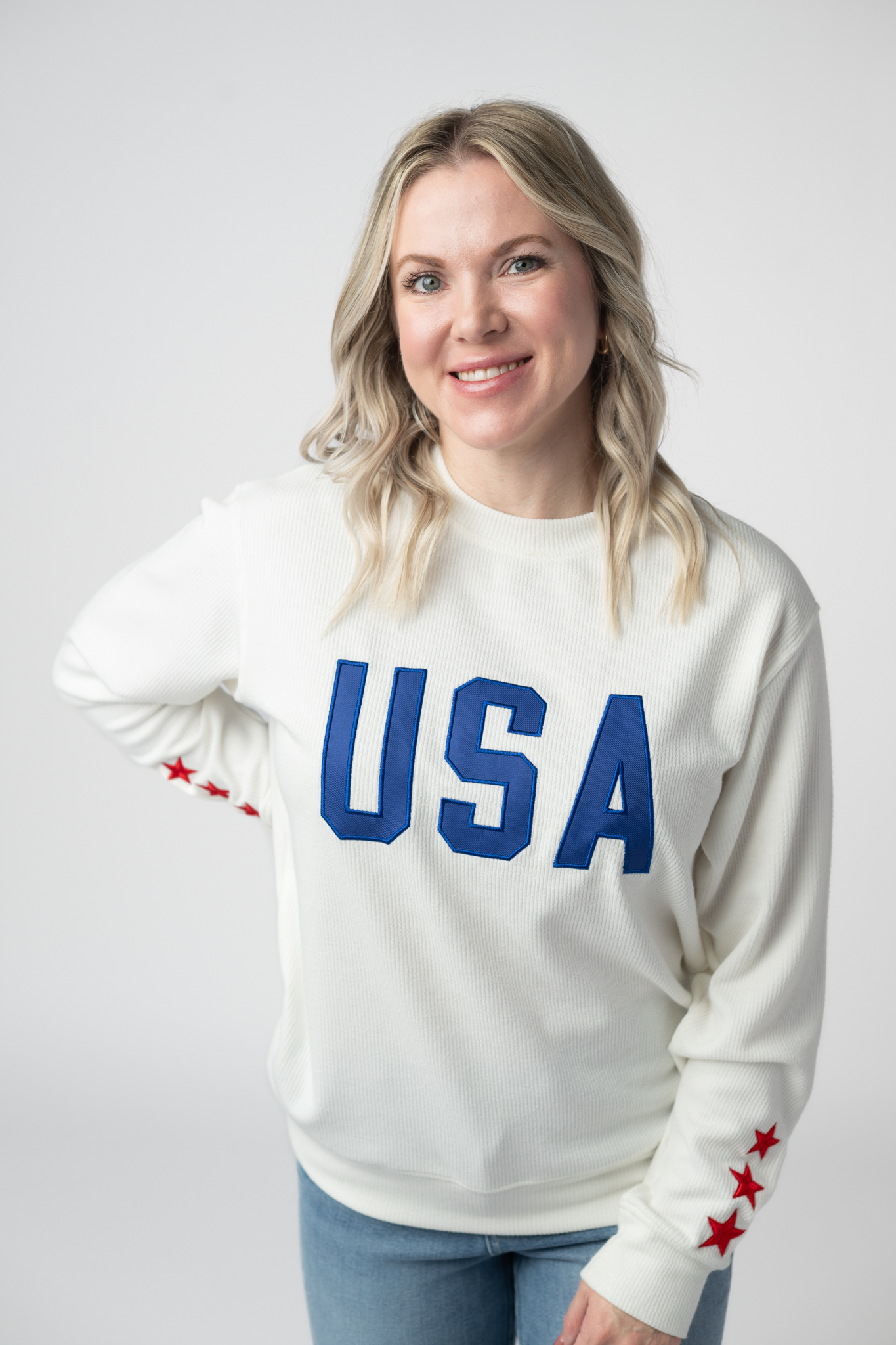 Woman wearing a white ribbed crew neck pullover sweatshirt with blue USA applique lettering on chest and red star accents on sleeves, paired with light wash jeans