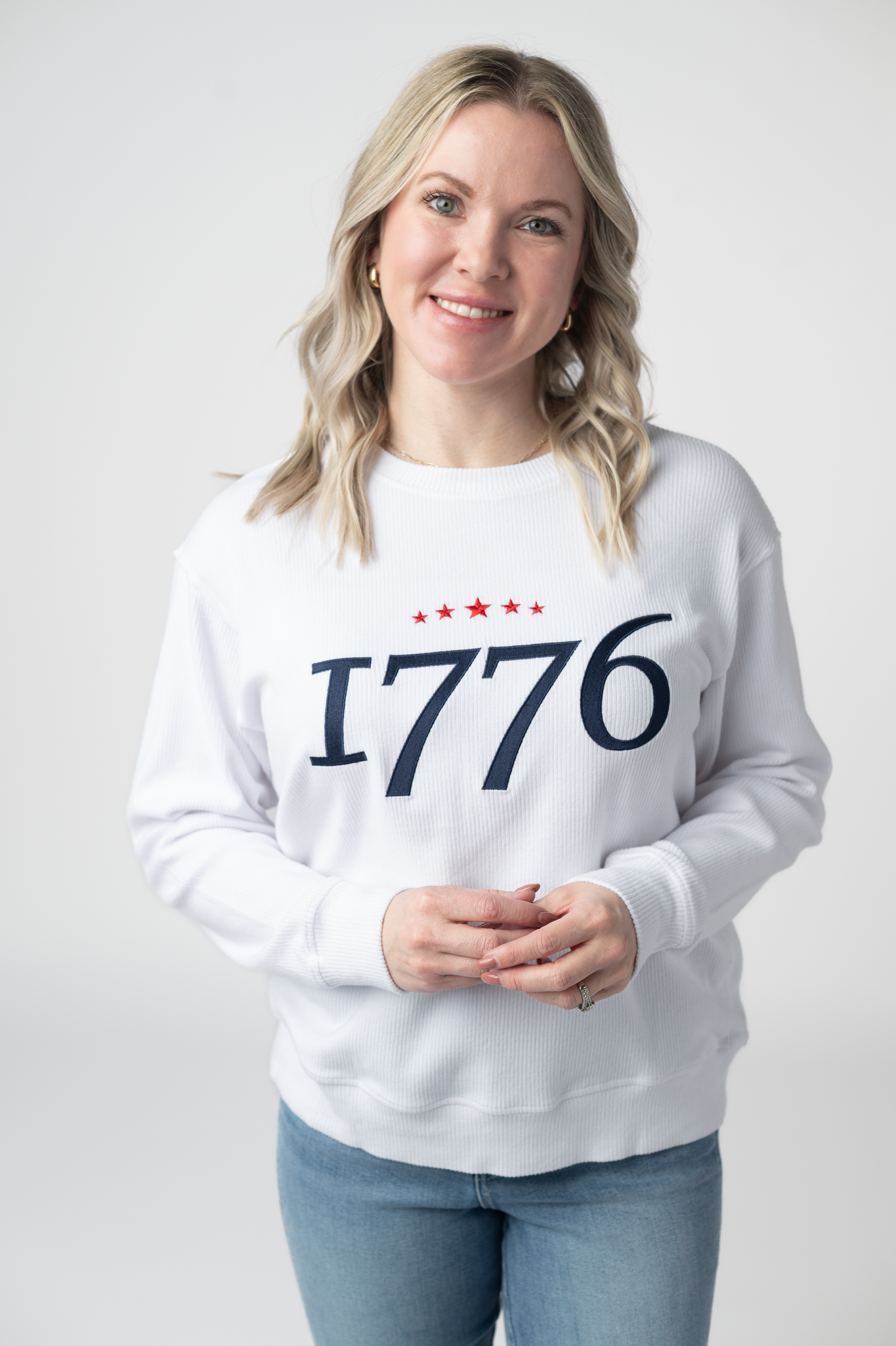Woman wearing a white ribbed crew neck pullover top with navy embroidered 1776 text and red star accents across the chest, paired with light wash jeans
