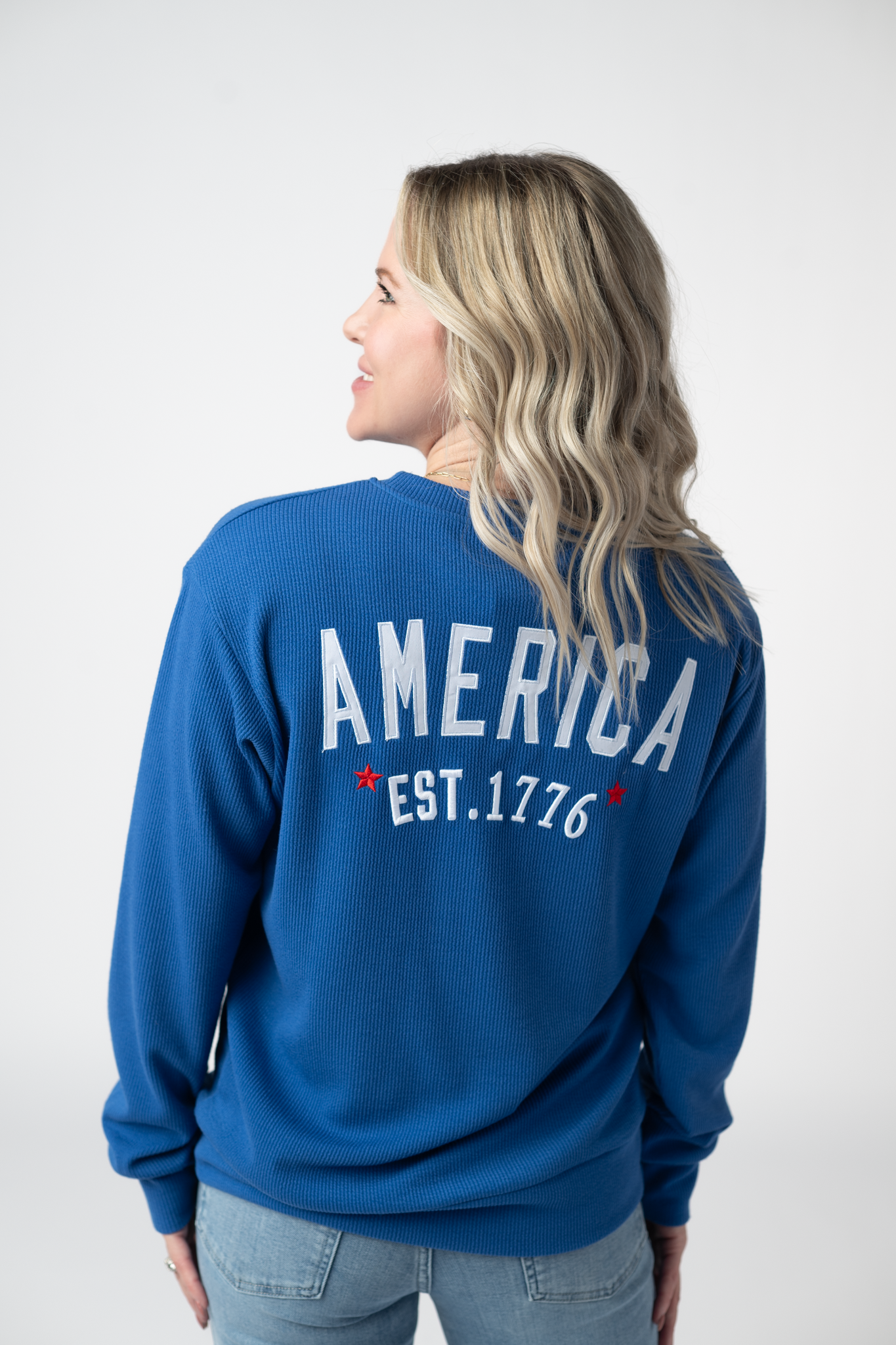 Woman wearing a blue America pullover sweatshirt with white embroidered America Est. 1776 text and red stars on the back, paired with light wash jeans